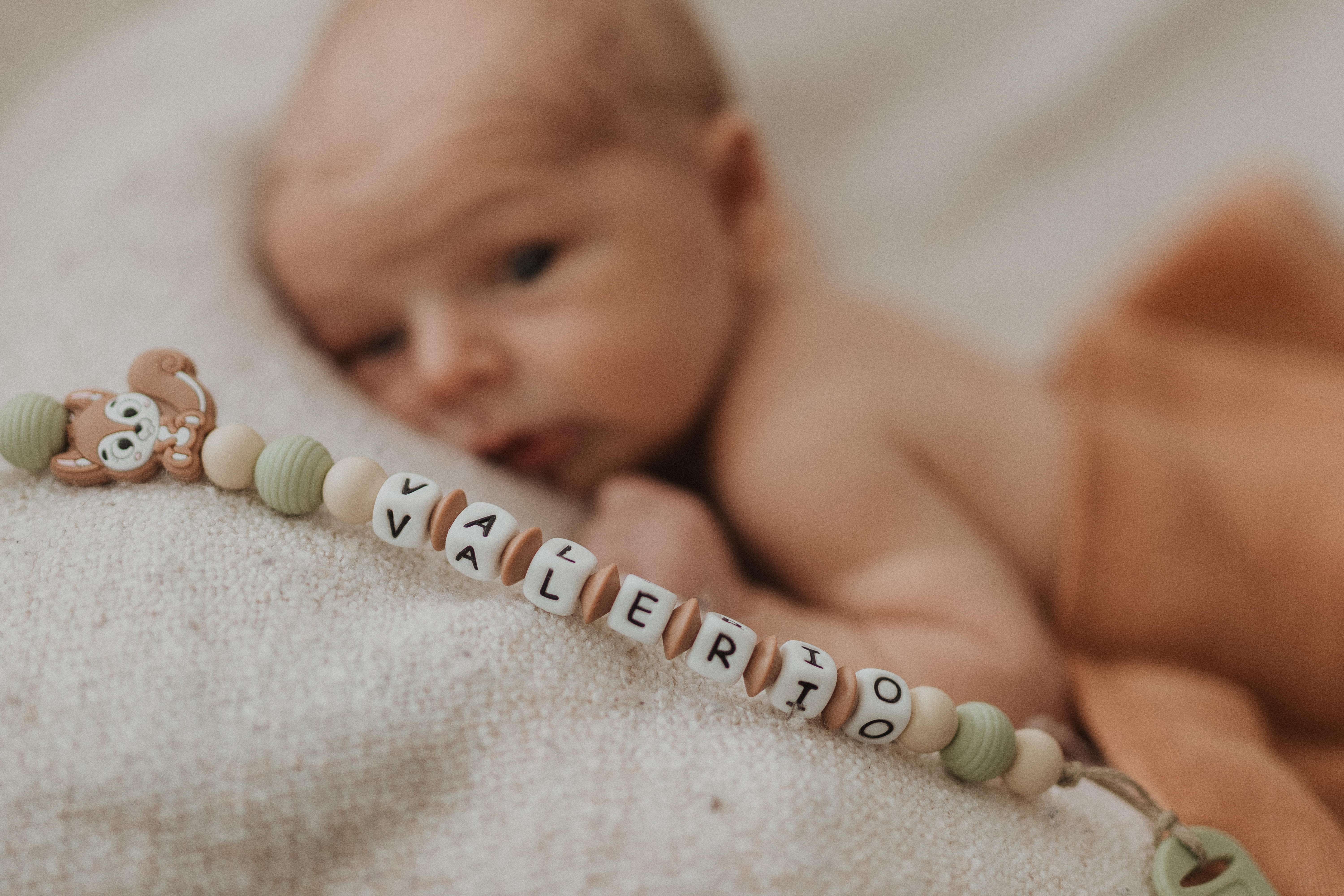 Eine schöne Detailaufnahme der Namenskette eines Babys namens Alessio. Hinter der Kette ist verschwommen das Gesicht des Kindes zu sehen. Eine spezielle Aufnahme während eines Fotoshootings für Neugeborene.
