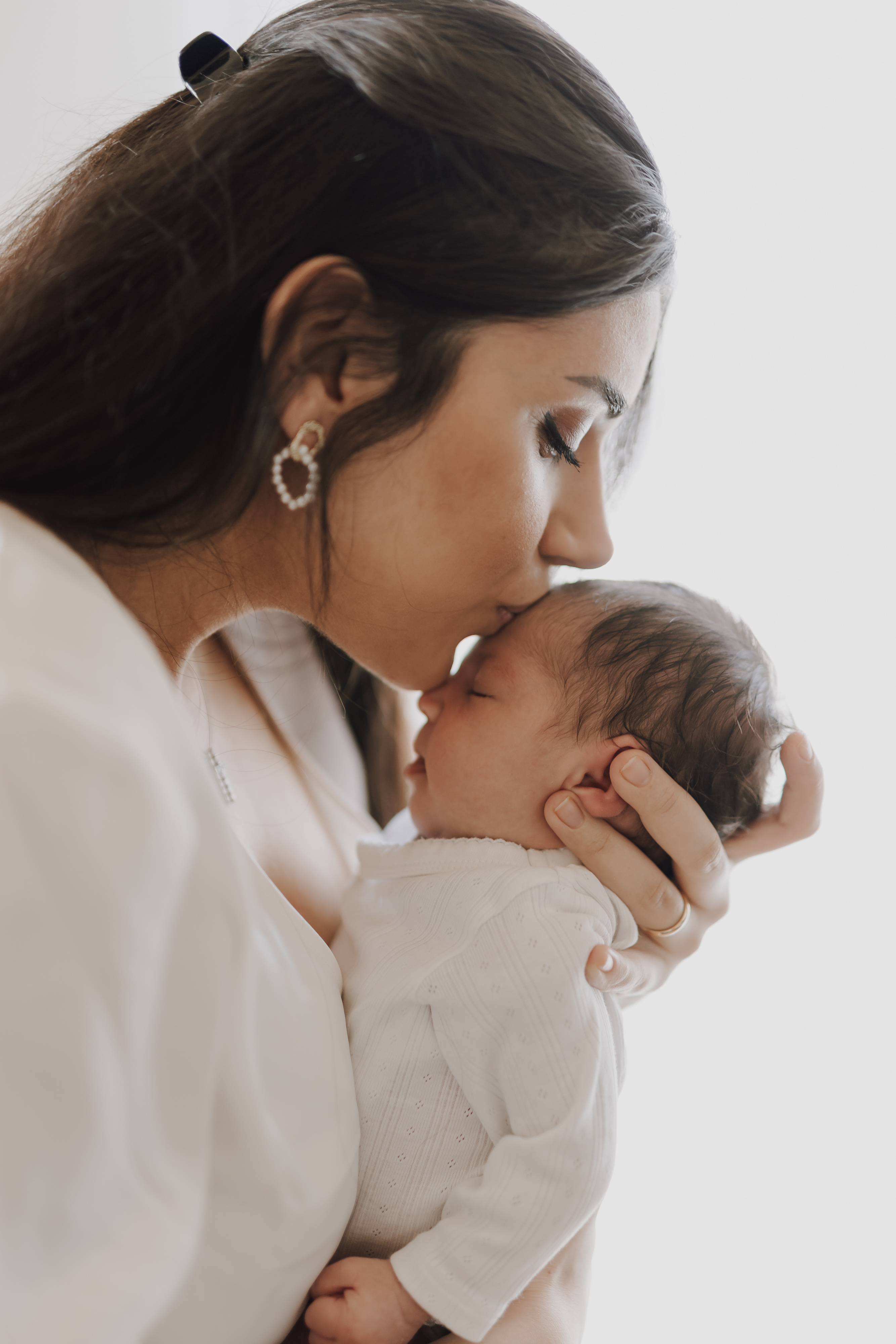 Eine Frau mit langen braunen Haaren und einem weißen Oberteil küsst die Stirn eines schlafenden Neugeborenen, das sie während eines intimen Hausbesuchs im Arm hält. Beide haben sanfte und heitere Ausdrücke und der Hintergrund ist verschwommen, was ihre Verbindung in diesem zarten Moment des Neugeborenenshootings betont.