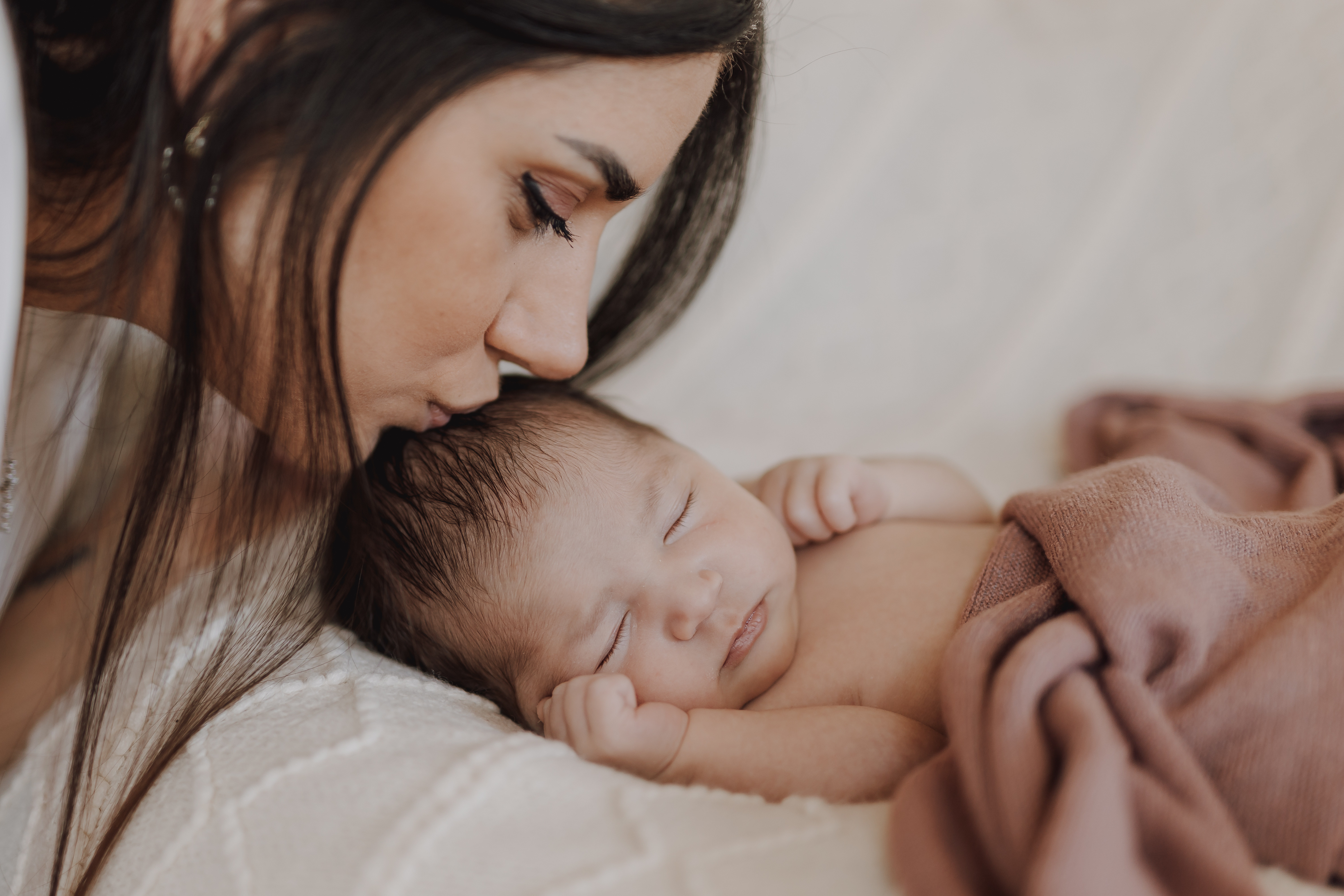 Eine Frau küsst sanft die Stirn eines schlafenden Babys, das in eine braune Decke gewickelt ist. Die ruhige und intime Umgebung, verstärkt durch sanftes Licht, fängt den zarten Moment ein. Diese wunderschöne Szene könnte das perfekte Neugeborenenfoto für jede Fotoshooting-Sitzung sein.
