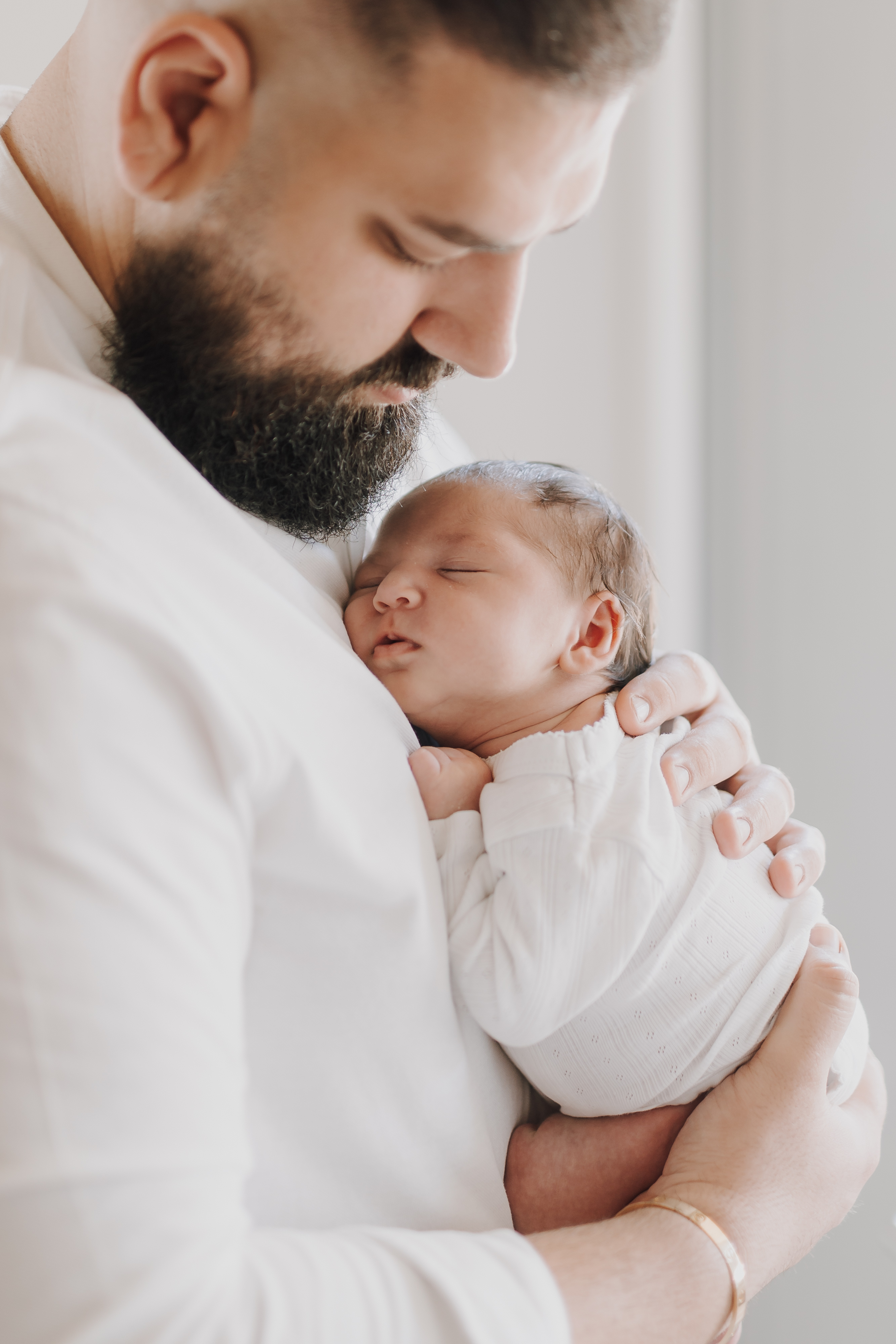 Ein bärtiger Mann wiegt ein schlafendes Baby sanft an seiner Brust, beide sind weiß gekleidet. Der Mann blickt zärtlich auf das Baby herab, von der Seite fällt sanftes Licht ein. Diese Newbornshooting-Szene vermittelt Wärme und Zuneigung.