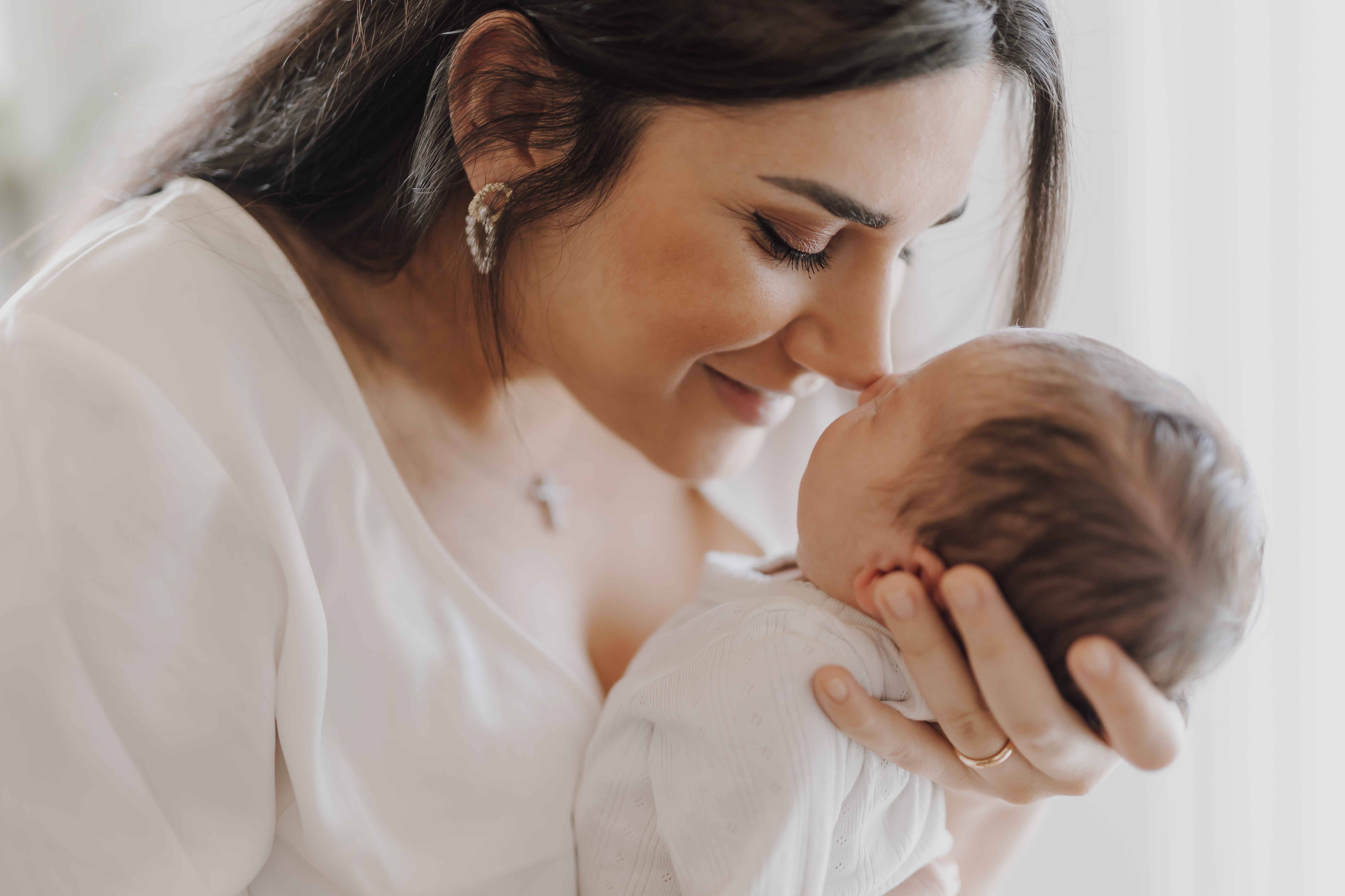 Eine Frau mit langen dunklen Haaren, Perlenohrringen und einem weißen Oberteil hält ihre Nase sanft an die Nase eines Neugeborenen und schmiegt sie an sie. Während des Fotoshootings hat sie einen zufriedenen, liebevollen Gesichtsausdruck. Das Baby wird im Rahmen des Hausbesuchs der Neugeborenenfotografin in eine weiße Decke gewickelt.