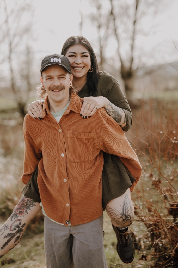 Ein Mann in einer braunen Cordjacke und einer schwarzen Mütze trägt eine Frau mit langen dunklen Haaren und einem grünen Kleid huckepack. Beide lächeln in der natürlichen Umgebung im Freien mit Bäumen und Sträuchern im Hintergrund und fangen während ihres fröhlichen Paarshootings die Essenz natürlicher Paarfotografie ein.