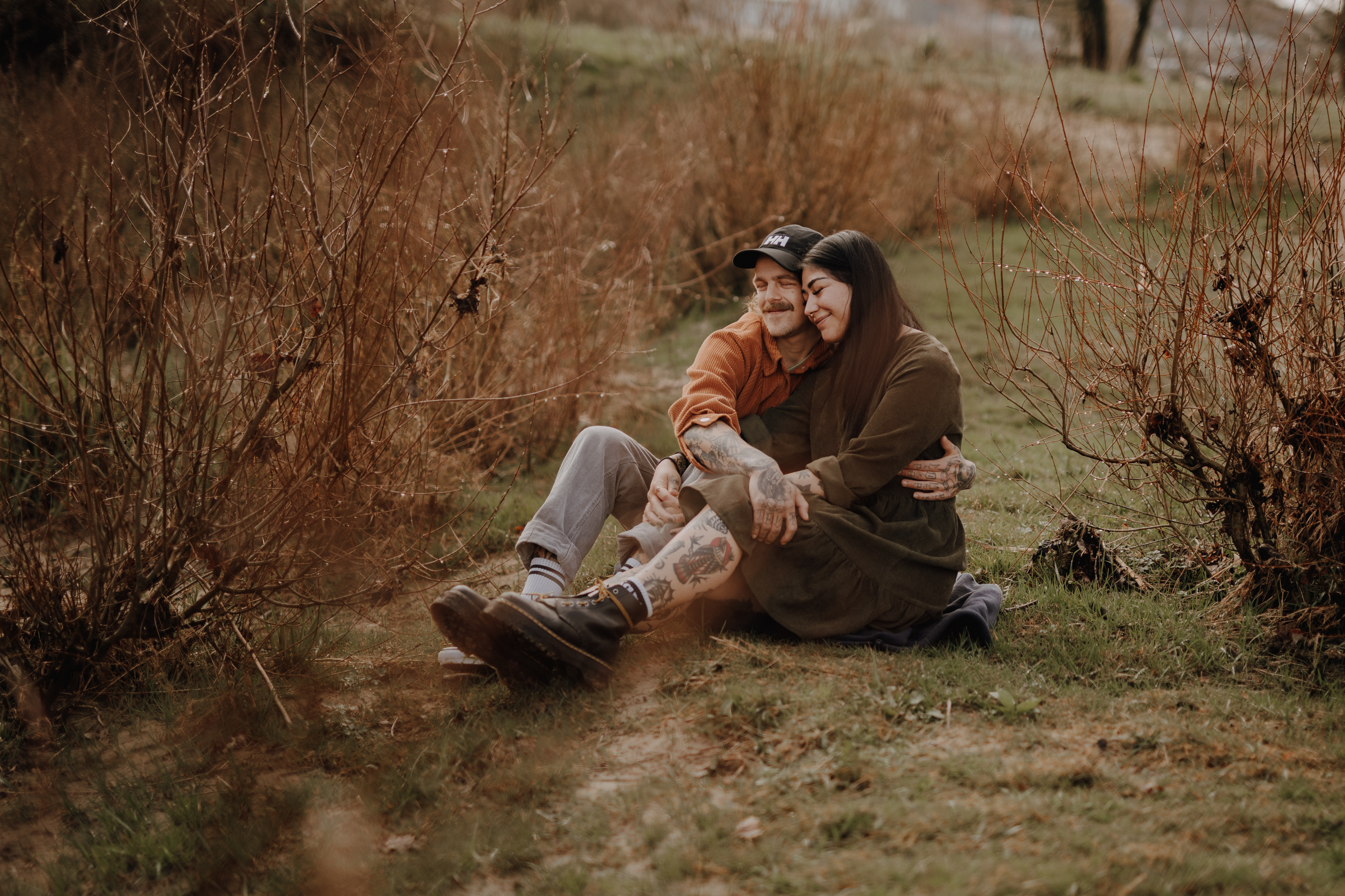 Ein Paar sitzt im Gras inmitten spärlicher, brauner Büsche und hält einen wunderschönen Moment in seiner Paarfotografie fest. Sie lächeln und umarmen sich ganz natürlich. Die Person links trägt einen Hut und eine orangefarbene Jacke, während die Person rechts ein braunes Kleid trägt. In dieser Outdoor-Fotoshooting-Kulisse wirken sie glücklich und entspannt.