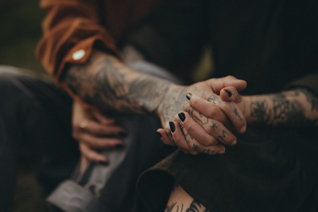 Nahaufnahme von zwei tätowierten Personen, die sich bei einem Paarshooting an den Händen halten. Die Person im Vordergrund hat schwarzen Nagellack und beide haben verschiedene Tattoos an Händen und Armen. Eine trägt eine dunkle Jacke und die andere einen hochgekrempelten Ärmel in Braun. Der Hintergrund ist verschwommen und fängt einen intimen Moment durch die Linse des Fotografen ein.