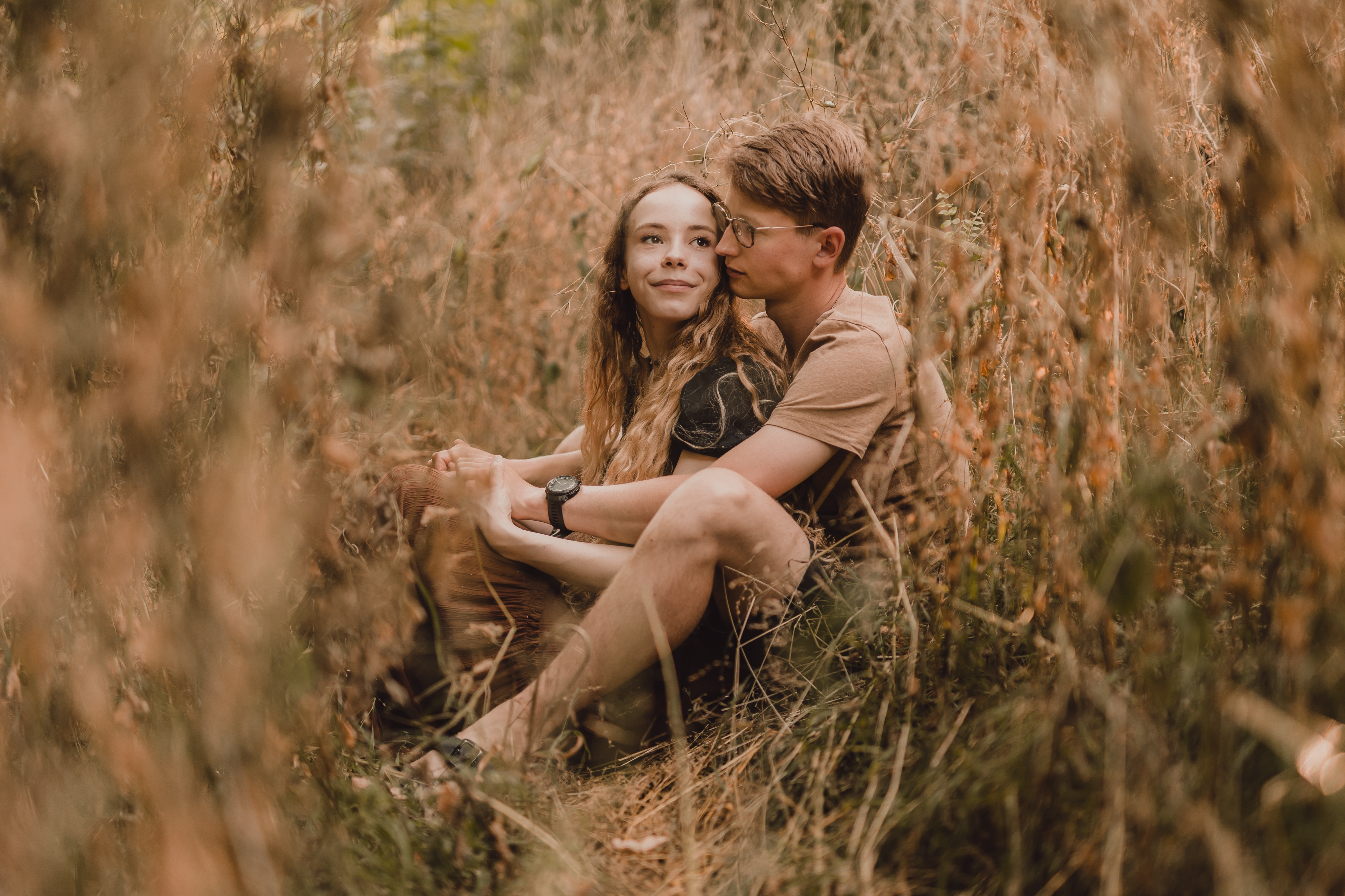 Ein junges Paar sitzt eng beieinander auf einem Feld aus hohem, trockenem Gras. Die Frau mit langen Haaren blickt zufrieden in die Ferne, während der Mann mit Brille und kurzen Haaren sie liebevoll ansieht. In dieser natürlich schönen Kulisse für ihr Paarshooting sind beide leger gekleidet und von herbstlichen Farbtönen umgeben.