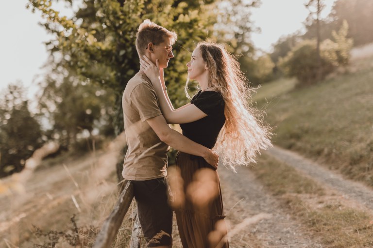 Ein Paar steht einander gegenüber auf einem sonnenbeschienenen Pfad auf einer Wiese, im Hintergrund sind Bäume zu sehen. Die Frau mit dem langen wallenden Haar hält sanft das Gesicht des Mannes, während er seine Hände auf ihre Taille legt. Sie blicken sich liebevoll in die Augen, perfekt für eine zeitlose Paarfotografie während ihres Fotoshootings.