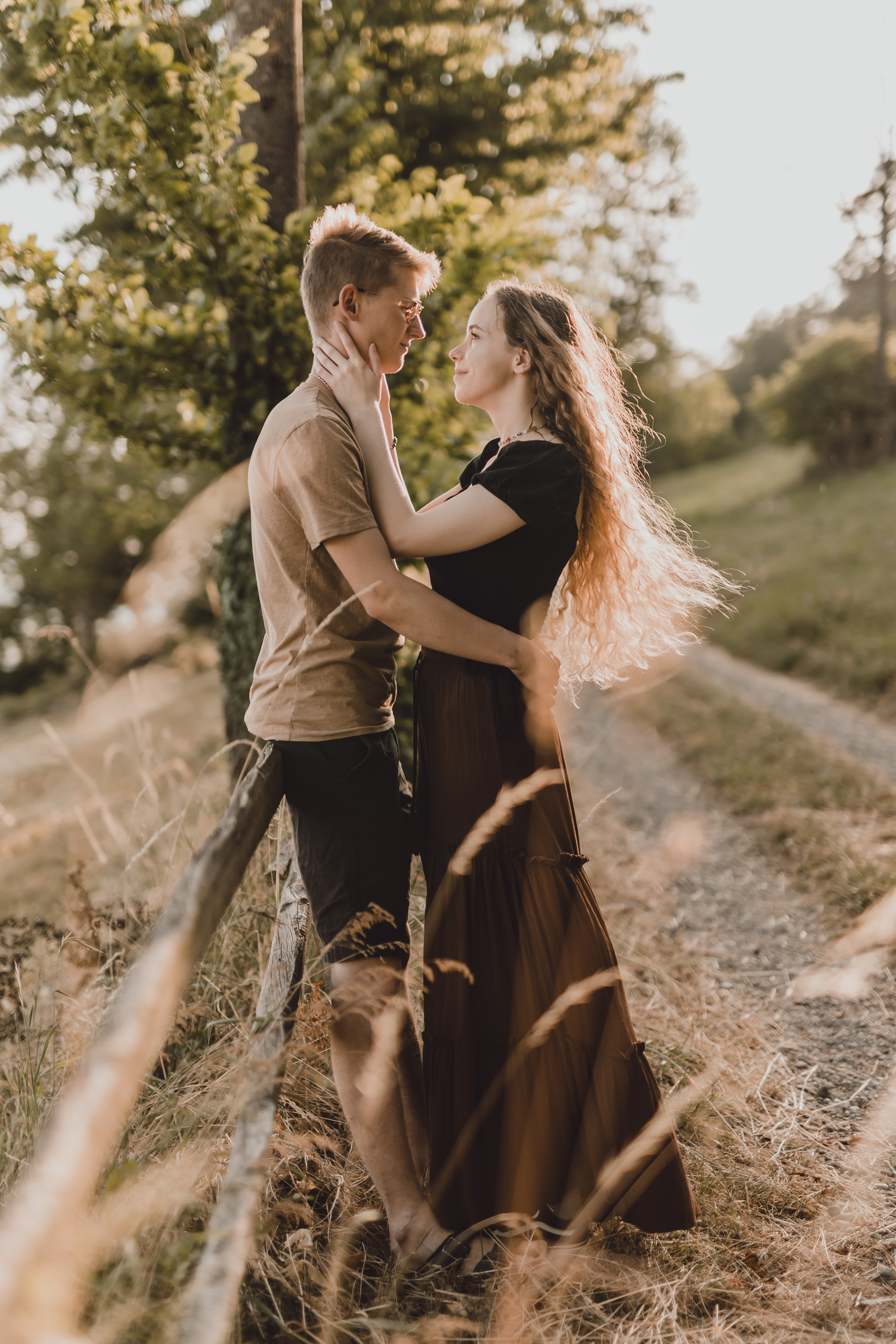 Ein Paar steht eng beieinander auf einer sonnenbeschienenen Wiese in der Nähe eines Holzzauns. Die Frau mit dem langen, wallenden Haar hält sanft das Gesicht des Mannes, und sie schauen sich in die Augen und teilen einen intimen Moment. Im Hintergrund sind Bäume und ein Pfad zu sehen, während der Fotograf diese natürliche Szene einfängt.