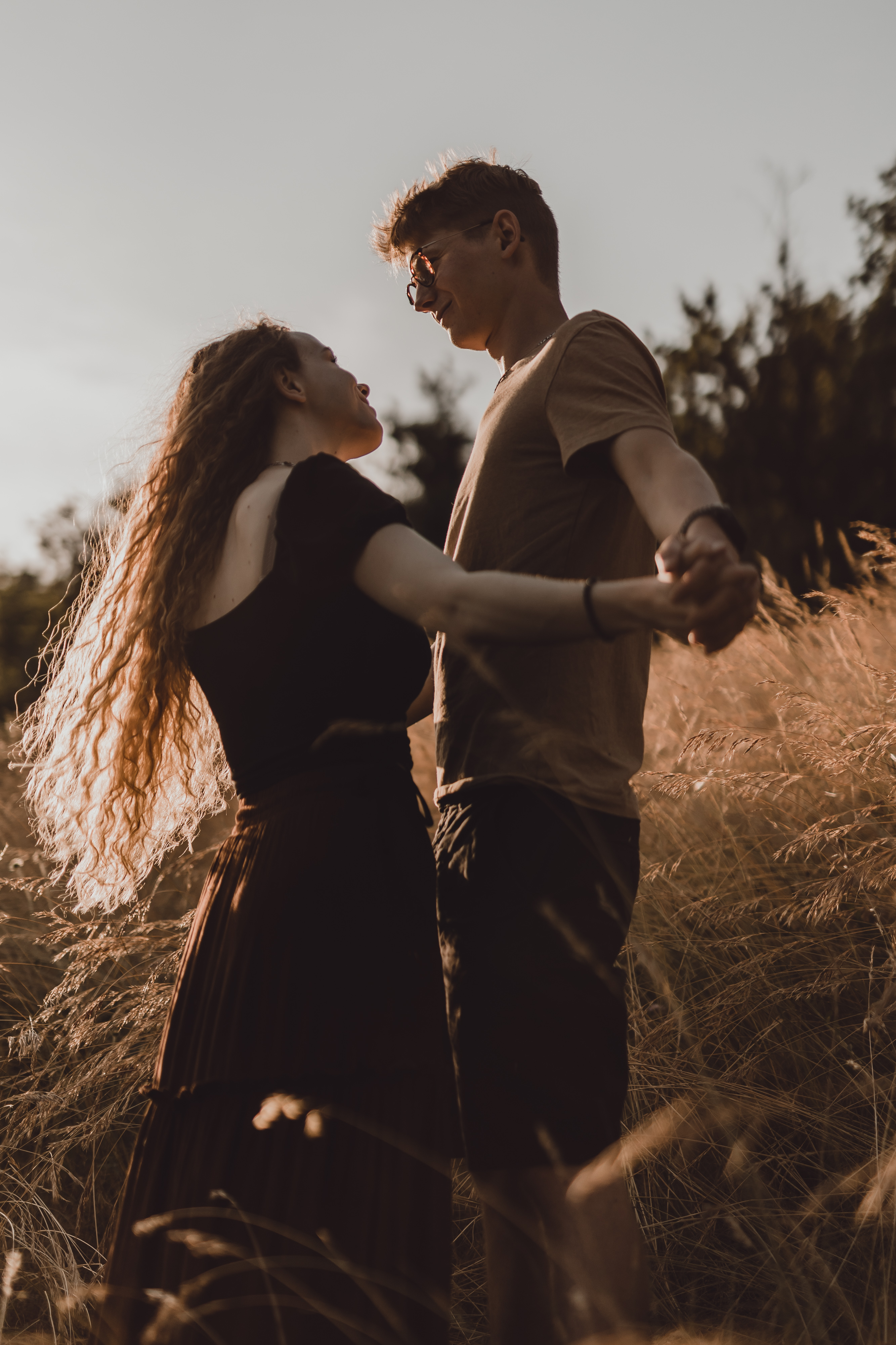 Ein Paar steht in einem sonnenbeschienenen Feld mit hohem Gras, hält sich an den Händen und schaut sich liebevoll an. Die Person links hat langes lockiges Haar und die Person rechts trägt eine Sonnenbrille. Die untergehende Sonne wirft einen warmen, goldenen Schein über die Paarfotografie-Szene.