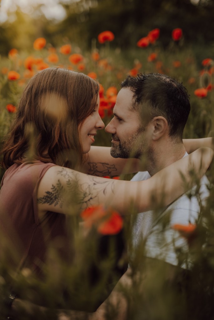 Ein Paar sitzt eng beieinander in einem Feld aus roten Blumen und fängt damit die Essenz der Paarfotografie ein. Sie stehen sich gegenüber, lächeln und berühren ihre Stirnen fast. Die Frau hat welliges rotes Haar und Tattoos auf dem Arm, während der Mann kurzes dunkles Haar und Stoppeln hat. Große, verschwommene Blumen umgeben sie.