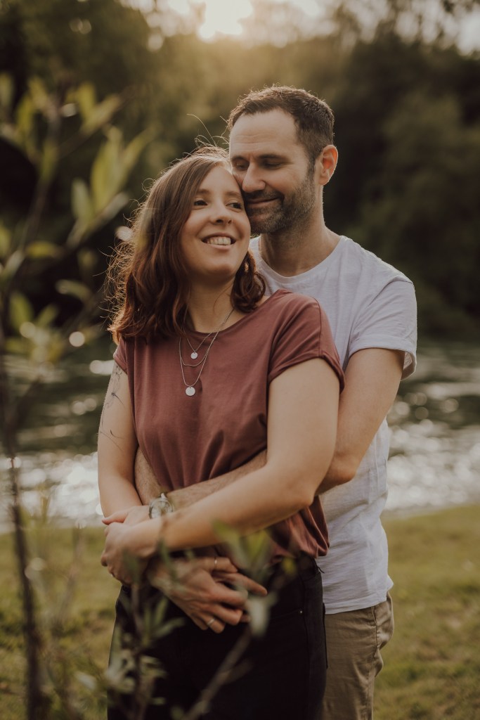 Eine lächelnde Frau mit schulterlangem braunem Haar steht vor einem Mann mit kurzen dunklen Haaren, der sie von hinten umarmt. Sie sind draußen in der Nähe eines Gewässers, umgeben von Grün, perfekt für eine romantische Paarfotografie-Session. Die Frau trägt ein kastanienbraunes Hemd und der Mann ist weiß gekleidet.