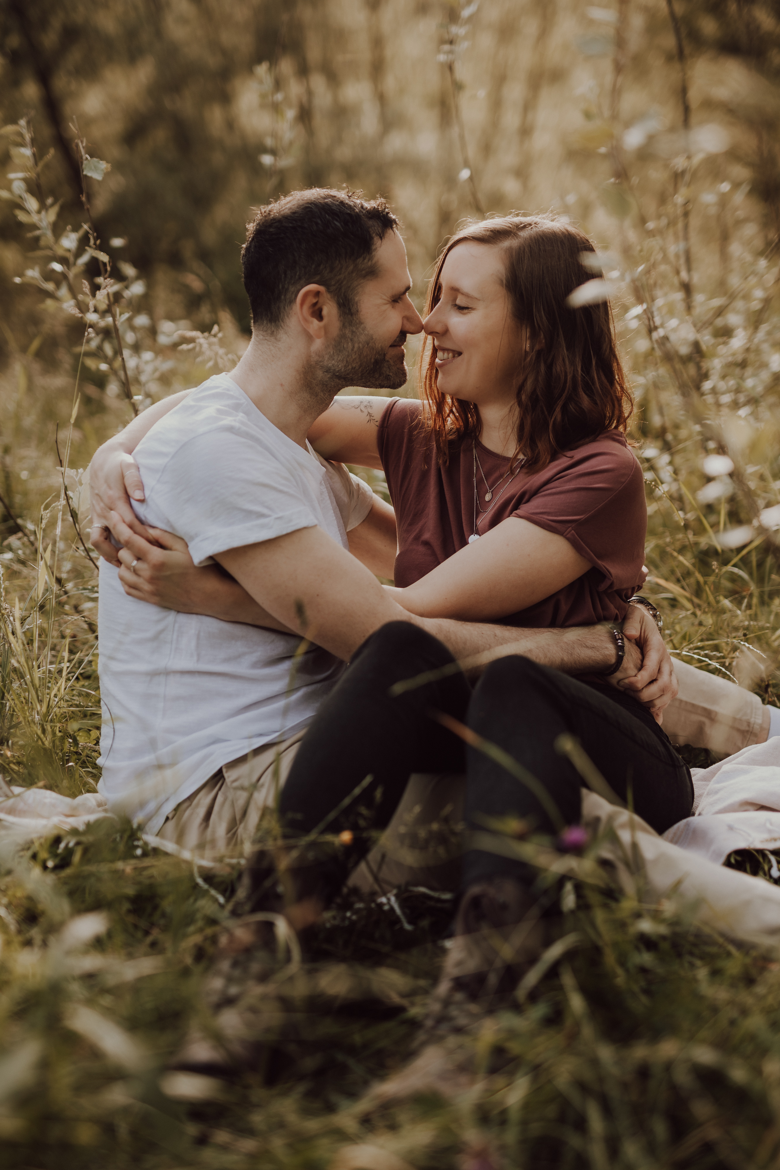 Ein Paar sitzt auf einer Wiese, umarmt sich und lächelt sich an. Der Mann trägt ein weißes T-Shirt und helle Hosen, während die Frau ein kastanienbraunes T-Shirt und schwarze Hosen trägt. Die Umgebung wirkt friedlich mit hohem Gras und sanftem, natürlichem Licht, perfekt für ein intimes Paarshooting.