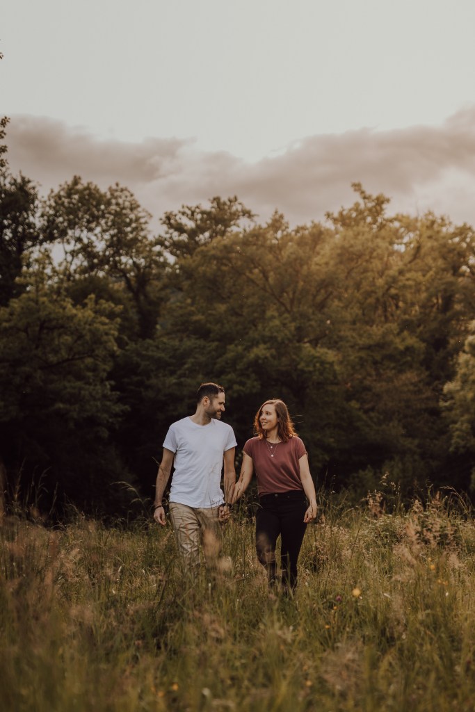 Ein Paar geht Hand in Hand durch eine Wiese. Der Mann trägt ein weißes T-Shirt und beige Shorts, während die Frau ein kastanienbraunes Oberteil und eine schwarze Hose trägt. Bäume und ein bewölkter Himmel bilden den Hintergrund, beleuchtet von sanftem, goldenem Licht – perfekt für ein natürliches Paarfotografie-Fotoshooting.