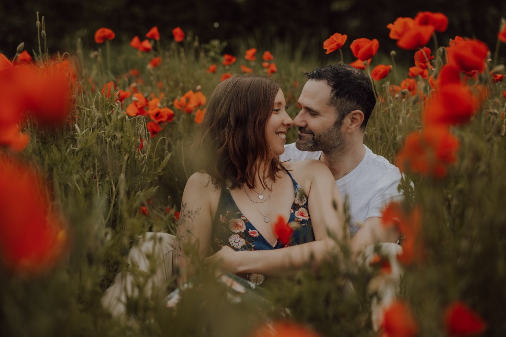 Ein Paar sitzt eng beieinander in einem Feld leuchtend roter Mohnblumen und blickt sich liebevoll an. Die Frau trägt ein geblümtes Kleid und der Mann ein helles Hemd. Das üppige Grün und die leuchtenden Blumen umgeben sie und schaffen eine romantische Kulisse, die perfekt für Paarfotografie ist.
