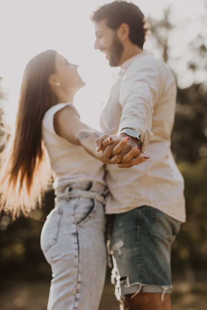 Ein Paar steht sich im Freien gegenüber und hält sich an den Händen. Die Frau mit langen Haaren, Jeans und weißem Oberteil, lehnt sich leicht zurück und lächelt. Der Mann mit kurzen Haaren und Bart, in einem weißen Hemd und Shorts aus Baden, lächelt ebenfalls. Sonnenlicht fällt in diesem natürlichen Moment der Paarfotografie durch die Bäume hinter ihnen.
