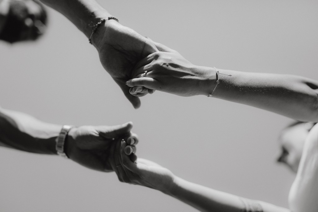 Ein Schwarzweißfoto fängt einen intimen Moment der Paarfotografie ein: Zwei Händepaare reichen sich einander, eines bietet etwas Kleines und Zylindrisches an, das andere streckt es aus, um es zu empfangen. Die Aufnahme aus niedriger Perspektive vor einem klaren Himmel verstärkt die Emotion dieser Paarshooting-Szene.
