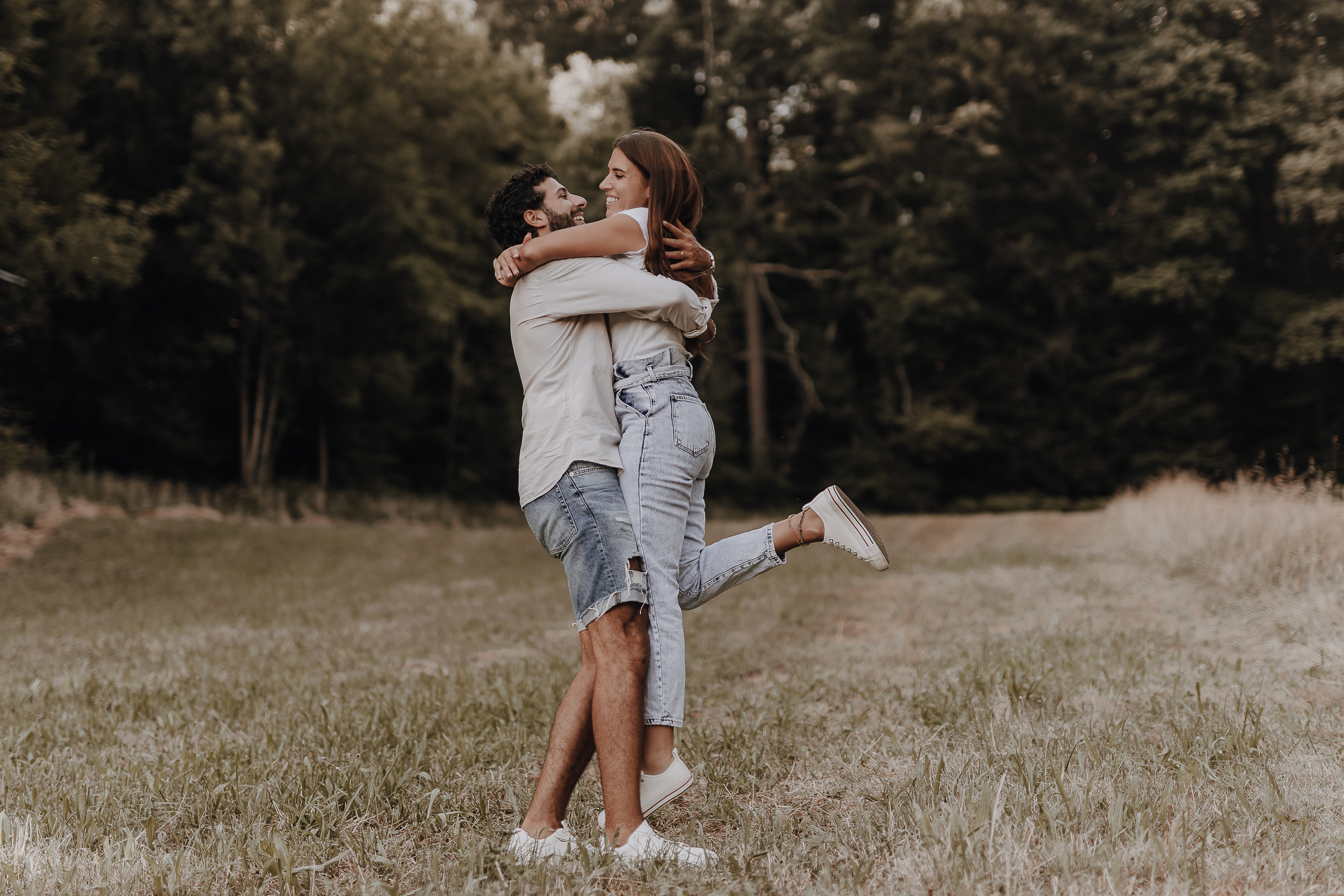 Ein Paar steht auf einem offenen Feld und umarmt sich beim Paarshooting. Die Frau hat ihre Arme um den Hals des Mannes gelegt und ein Bein vom Boden abgehoben, während der Mann sie stützt, indem er sie um die Taille hält. Beide tragen Freizeitkleidung und im Hintergrund sind grüne Bäume zu sehen.
