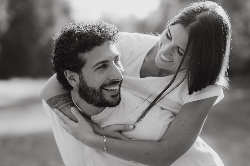 Ein Schwarz-Weiß-Foto eines liebevollen Paares im Freien während eines Paarshootings. Der Mann trägt die Frau Huckepack; beide lächeln und schauen sich an. Sie hat ihre Arme um seine Schultern gelegt und er hält ihre Beine und fängt einen freudigen und verspielten Moment in ihrer Paarfotografie ein.