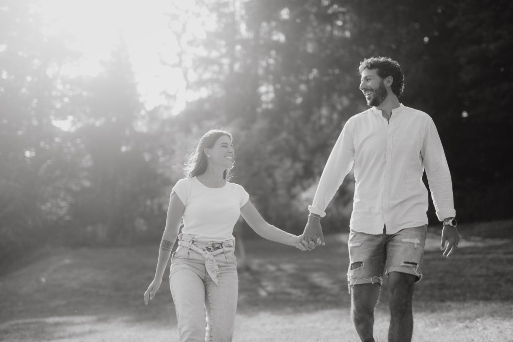 Schwarzweißbild eines Paares, das Hand in Hand im Freien mit Bäumen im Hintergrund spazieren geht. Beide lächeln und scheinen den Moment zu genießen. In natürlicher Paarfotografie trägt die Frau ein T-Shirt und Jeans, während der Mann ein langärmeliges Hemd und Shorts trägt.