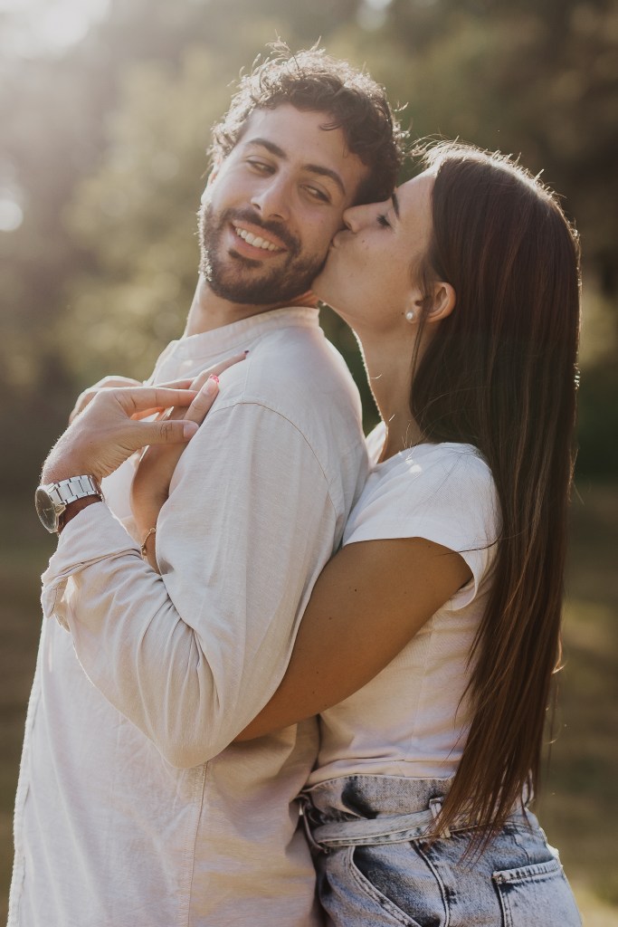 Eine Frau gibt ihrem Partner einen Kuss auf die Wange. sie umarmt ihn dabei von hinten, während er sein Gesicht zu ihr dreht und lacht. Echte Emotionen, eingefangen von der Paarfotografin Tasha Ender.
