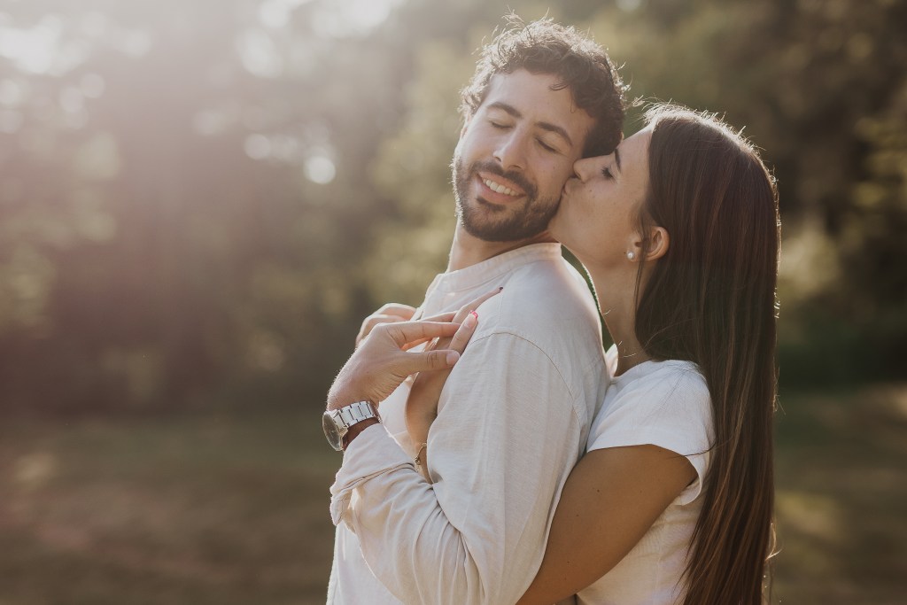 Eine Frau mit langen, braunen Haaren küsst die Wange eines Mannes mit lockigem Haar und Bart. Beide tragen helle Kleidung und lächeln mit geschlossenen Augen. Sie befinden sich draußen in einer sonnenbeschienenen, natürlichen Umgebung und genießen ein herrliches Paarshooting in Baden inmitten üppigen Grüns und sanften Sonnenlichts.