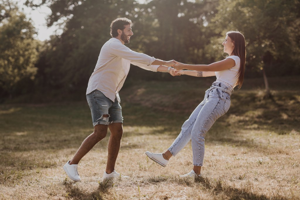 Ein Paar tanzt lachend im Sonnenschein des Sonnenuntergangs über eine Wiese. Momentaufnahme aus einem Paarshooting.