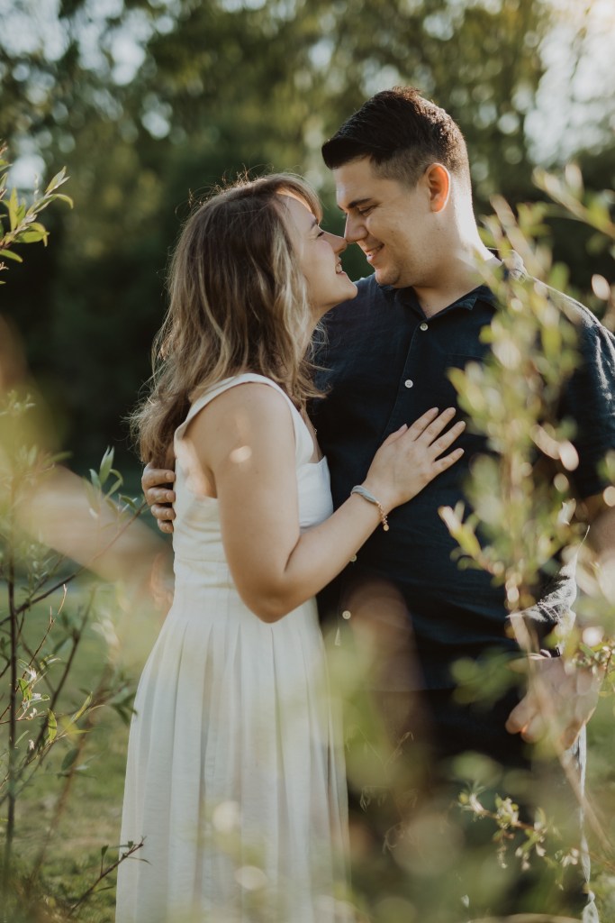 Ein Paar steht eng beieinander in einem Garten und lächelt sich liebevoll an. Die Frau im weißen Kleid hat ihre Hand auf der Brust des Mannes. Der Mann, der ein dunkles Hemd trägt, hat seinen Arm um sie gelegt. Sonnenlicht fällt durch das Grün und wirft einen warmen Schein – solch natürliche Paarfotografie fängt ihre Liebe wunderschön ein.