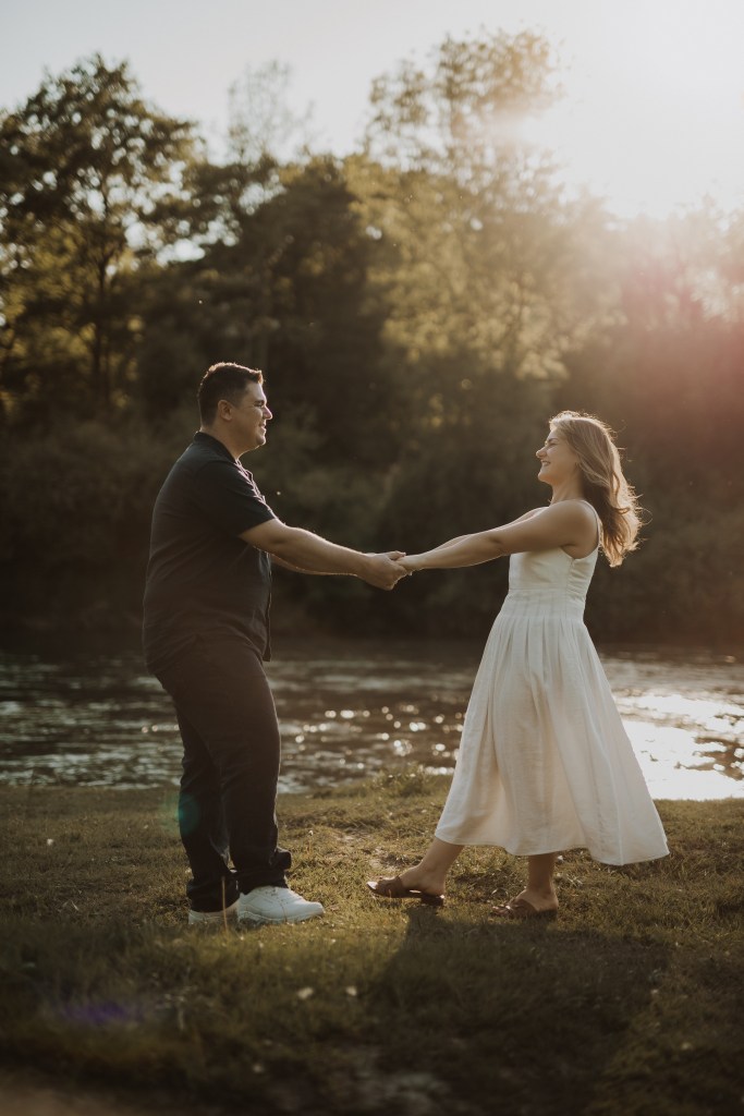 Ein Paar hält sich an den Händen und blickt sich lächelnd in einer sonnigen Umgebung im Freien in der Nähe eines Flusses an. Der Mann trägt ein schwarzes Hemd und eine schwarze Hose und die Frau ein weißes Kleid. Im Hintergrund sind üppige grüne Bäume und ein ruhiger Fluss zu sehen, was es zu einem idealen Ort für ein Fotoshooting für einen Fotografen macht.