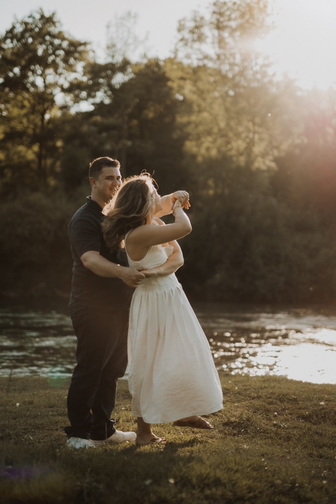Ein Paar umarmt sich auf einer Wiese in der Nähe eines Gewässers, getaucht in warmes, goldenes Sonnenlicht. Die Frau, die ein fließendes weißes Kleid trägt, steht vor ihr, während der Mann, in dunklem Hemd und Hose, während ihres Paarshootings hinter ihr steht. Im Hintergrund sind Bäume zu sehen, während sie den Moment genießen.