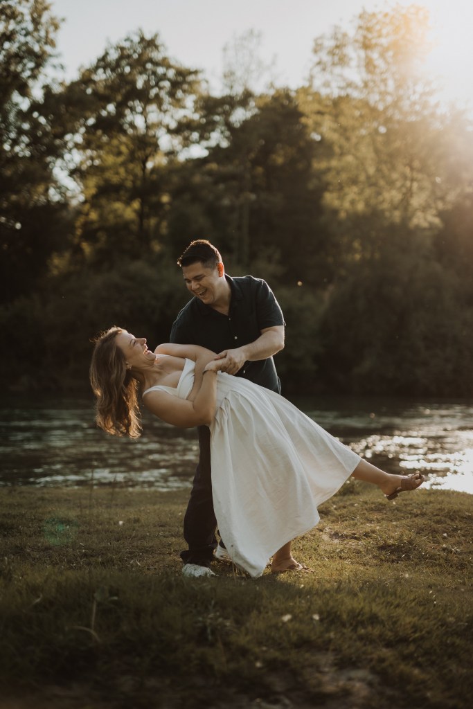 Ein Paar tanzt im Freien neben einem Fluss mit Bäumen im Hintergrund. Die Frau im weißen Kleid wird spielerisch von dem Mann im dunklen Hemd umarmt. Sonnenlicht fällt durch die Bäume und verleiht der Szene einen warmen, goldenen Schimmer – perfekt für jeden Fotografen, der die natürlichen Momente des Lebens festhält.