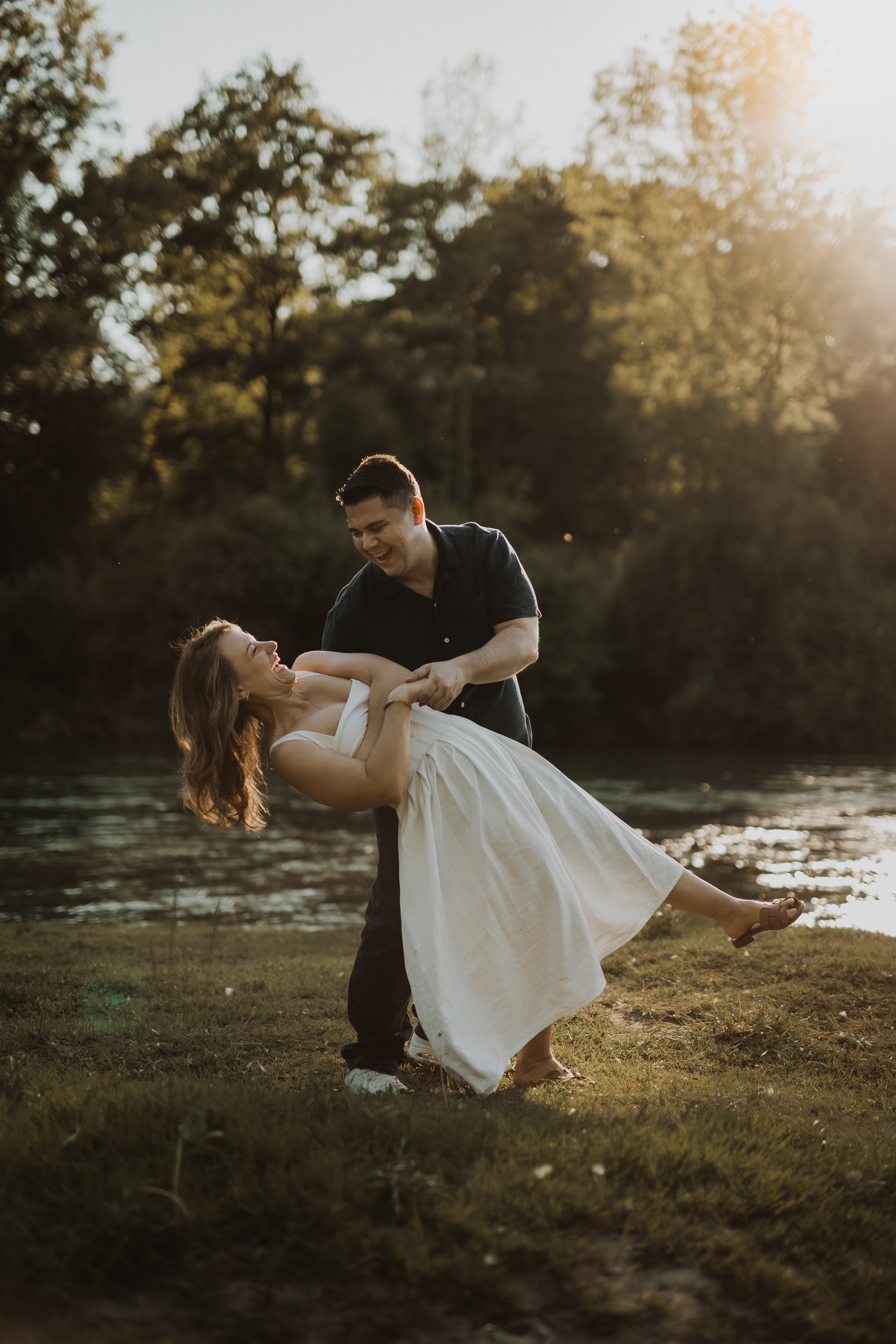 Ein Paar tanzt im Freien neben einem Fluss mit Bäumen im Hintergrund. Die Frau im weißen Kleid wird spielerisch von dem Mann im dunklen Hemd umarmt. Sonnenlicht fällt durch die Bäume und verleiht der Szene einen warmen, goldenen Schimmer – perfekt für jeden Fotografen, der die natürlichen Momente des Lebens festhält.