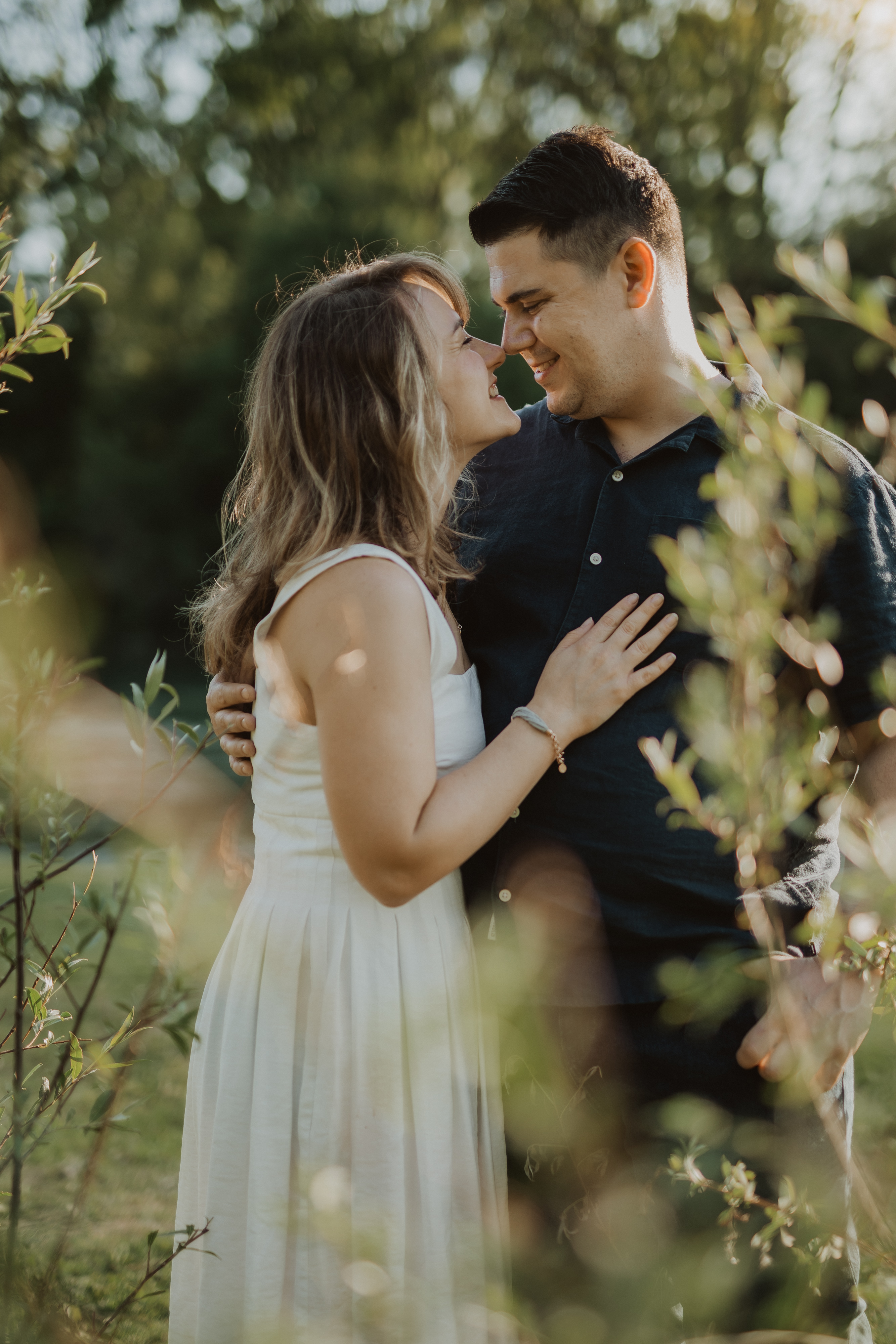Ein Paar steht draußen und blickt sich während eines Fotoshootings liebevoll an. Die Frau im weißen Kleid legt ihre Hand auf die Brust des Mannes, der ein dunkles Hemd trägt. Sie sind von Grün umgeben, durch dessen Blätter Sonnenlicht fällt und eine ruhige Atmosphäre schafft, die perfekt für Paarfotografie ist.