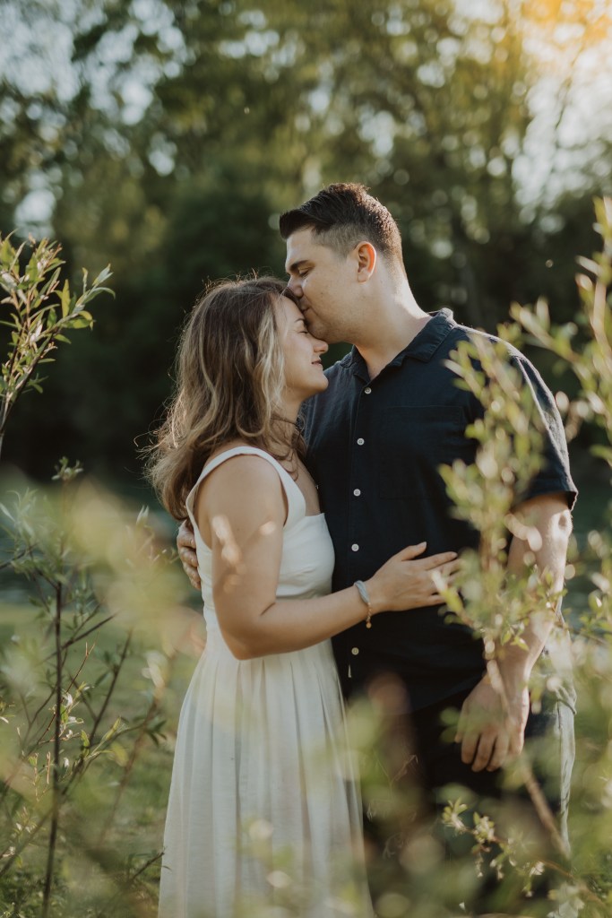 Ein Mann und eine Frau stehen eng beieinander in einer sonnenbeschienenen Außenkulisse, umgeben von Grün. Der Mann trägt ein dunkles Hemd und die Frau ein weißes Kleid. Während ihres Paarshootings küsst der Mann die Stirn der Frau, während sie sich umarmen und lächeln und einen zärtlichen Moment miteinander genießen.