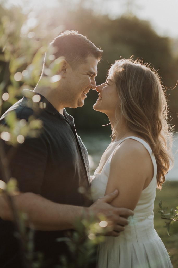 Ein Paar steht dicht beieinander in einer grünen Umgebung im Freien, ihre Nasen berühren sich und sie lächeln. Die Frau in einem ärmellosen weißen Kleid und der Mann in einem dunklen Hemd genießen das warme Sonnenlicht, das durch die Blätter um sie herum fällt. Dieser natürliche Moment wurde von einem talentierten Fotografen elegant festgehalten.