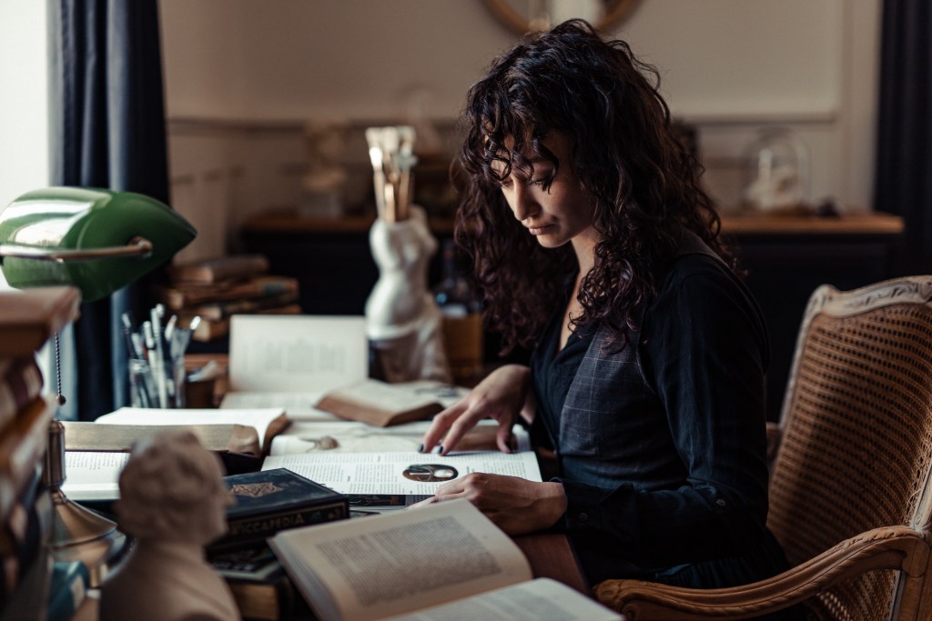 Eine Frau mit lockigem Haar sitzt an einem Holzschreibtisch, umgeben von offenen Büchern. Sie liest konzentriert, die Lupe in der Hand. Der gemütliche, warm beleuchtete Raum verfügt über eine grüne Schreibtischlampe und eine klassische Statue im Hintergrund – eine ideale Szene für ein Portraitshooting eines jeden erfahrenen Fotografen.