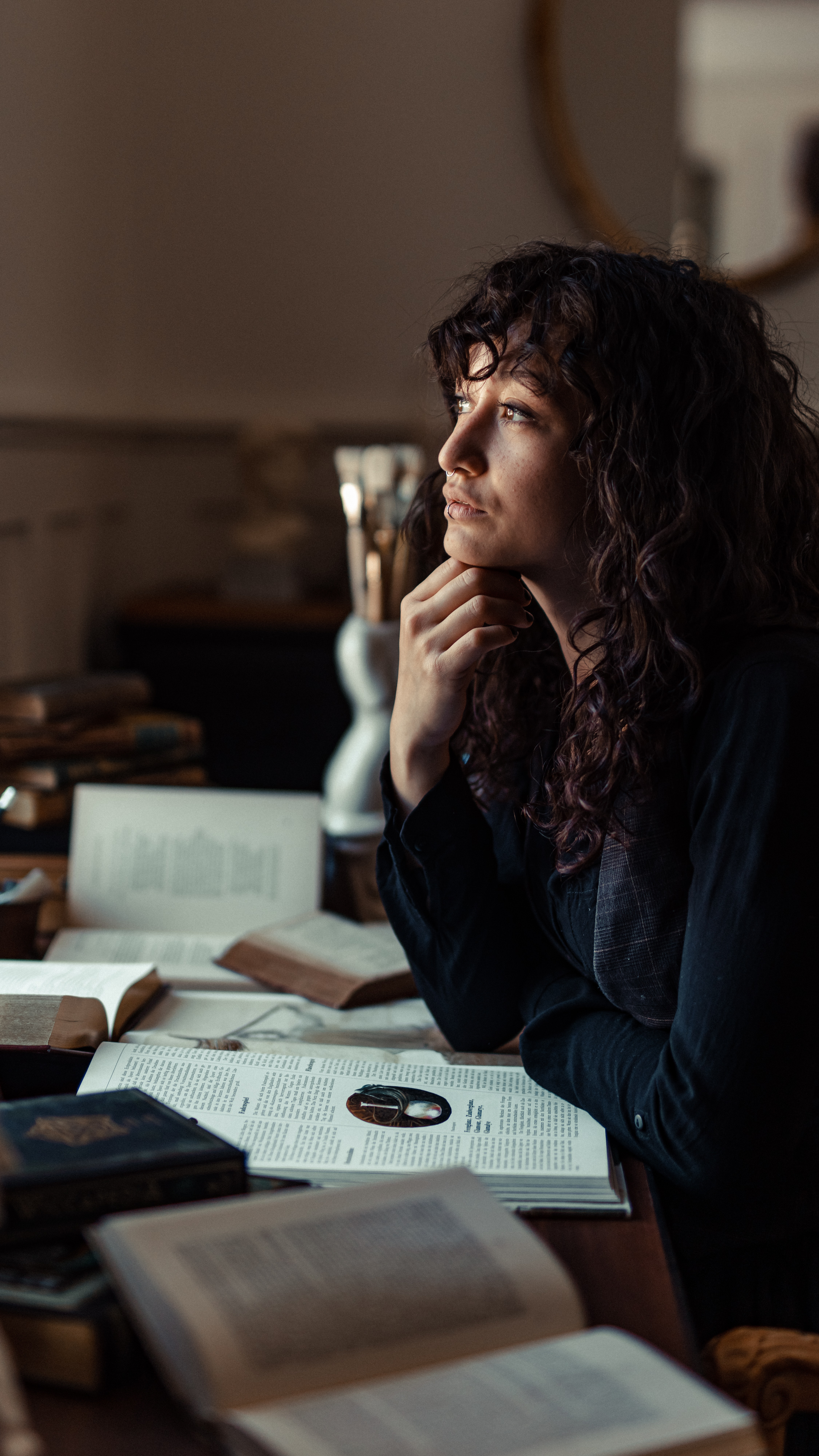 Eine junge Frau mit lockigem Haar sitzt an einem vollgestellten Holzschreibtisch und blickt nachdenklich aus dem Fenster. Sie stützt ihr Kinn auf ihre Hand, umgeben von offenen Büchern und einer alten Schreibmaschine, was eine natürlich gemütliche und besinnliche Atmosphäre schafft, die perfekt für ein Portrait-Fotoshooting ist.