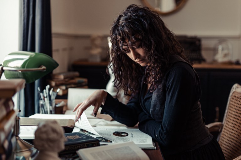 Eine Person mit lockigem Haar liest und blättert in einem Buch an einem überladenen Schreibtisch, vertieft in ihr Arbeitszimmer. Der Schreibtisch, bedeckt mit Büchern, Papieren und einer grünen Schreibtischlampe, schafft eine intellektuelle Atmosphäre. Im Hintergrund des Büros sind eine Schreibmaschine und andere Gegenstände zu sehen, die dieser intimen Porträtszene Tiefe verleihen.