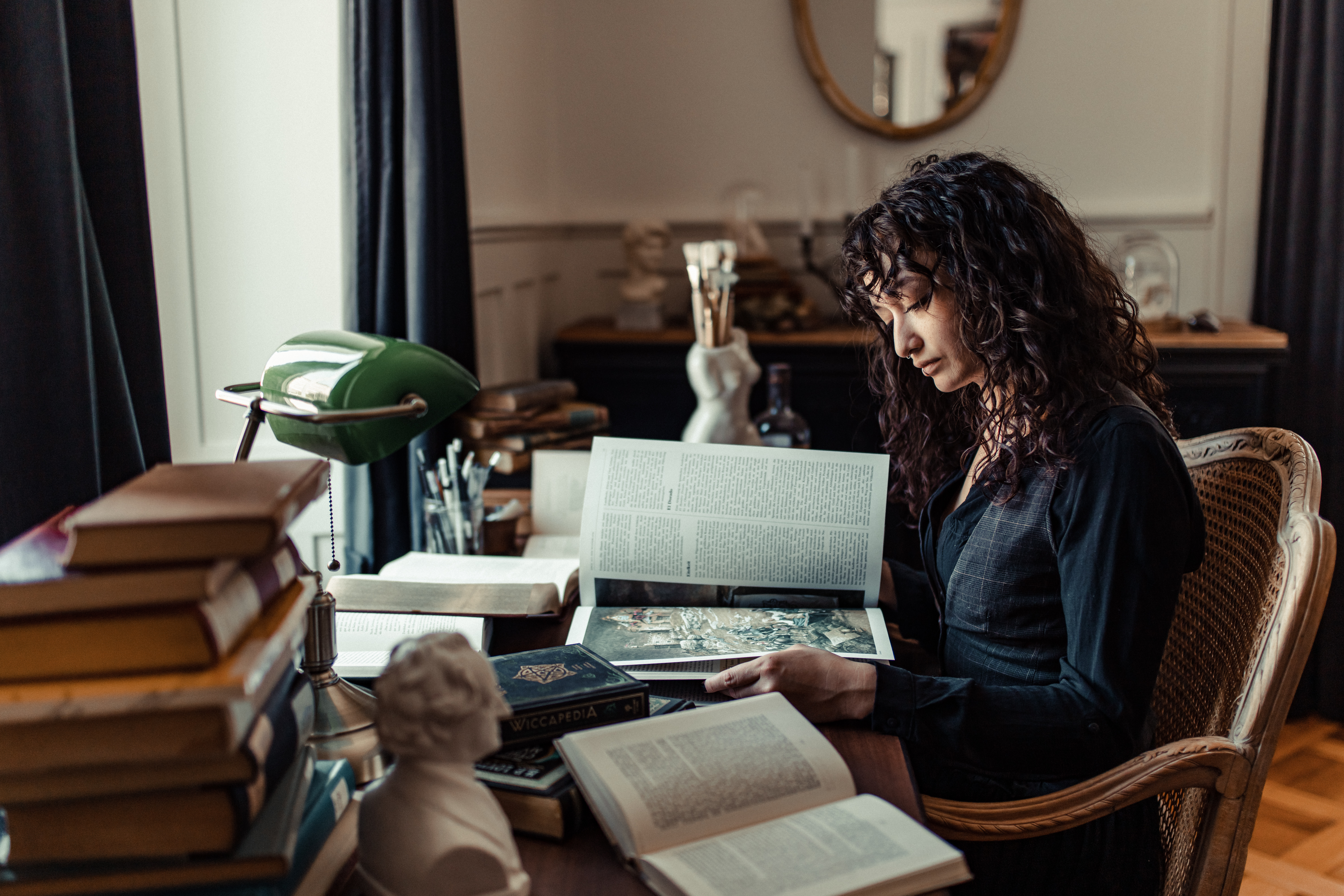 Eine Frau mit lockigem Haar sitzt in einem gemütlichen Zimmer an einem Holzschreibtisch und liest ein großes Buch. Der Schreibtisch ist mit Bücherstapeln und einer kleinen Statue vollgestopft und bietet so eine ideale Kulisse für ein Porträt. Eine grüne Schreibtischlampe beleuchtet den Arbeitsbereich, während dunkle Vorhänge ein Fenster im Hintergrund einrahmen.