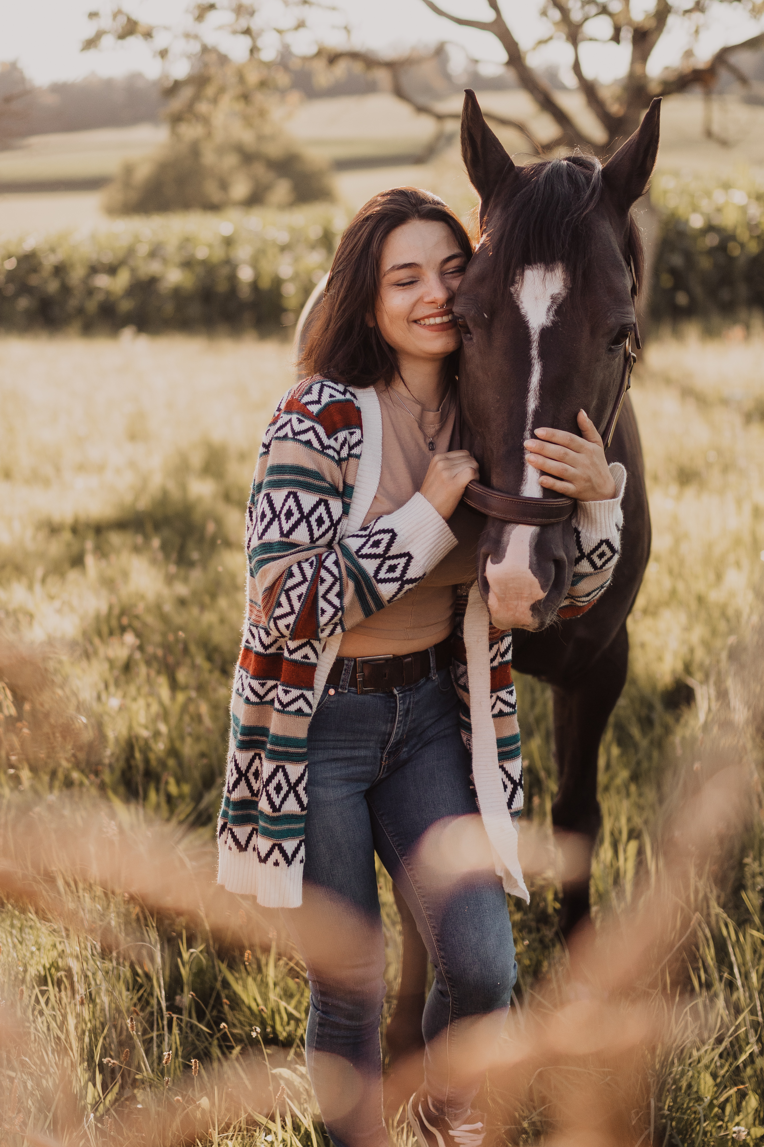 Eine Frau steht lächelnd auf einer sonnenbeschienenen Wiese und umarmt ein dunkelbraunes Pferd mit einer weißen Blesse im Gesicht. Sie trägt eine bunte, gemusterte Strickjacke über einem hellen Oberteil und Jeans. Im Hintergrund sind hohes Gras und ein Baum zu sehen, was die perfekte Kulisse für das Fotoshooting des Tierfotografen bildet.