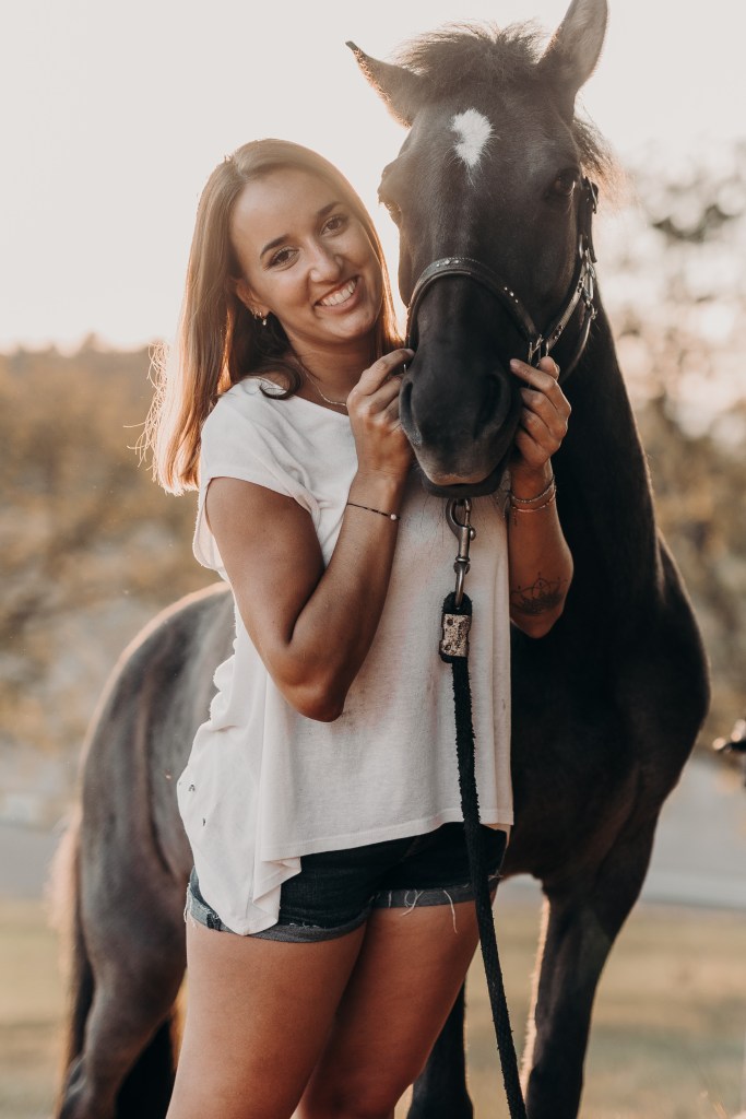 Eine lächelnde Frau in weißem Hemd und Jeansshorts steht neben einem schwarzen Pferd und hält mit beiden Händen das Zaumzeug. Die Außenkulisse mit ihrem unscharfen natürlichen Hintergrund ist perfekt für diese Pferdefotografie-Session. Das sanfte Sonnenlicht hebt die Gesichter beider wunderschön hervor.