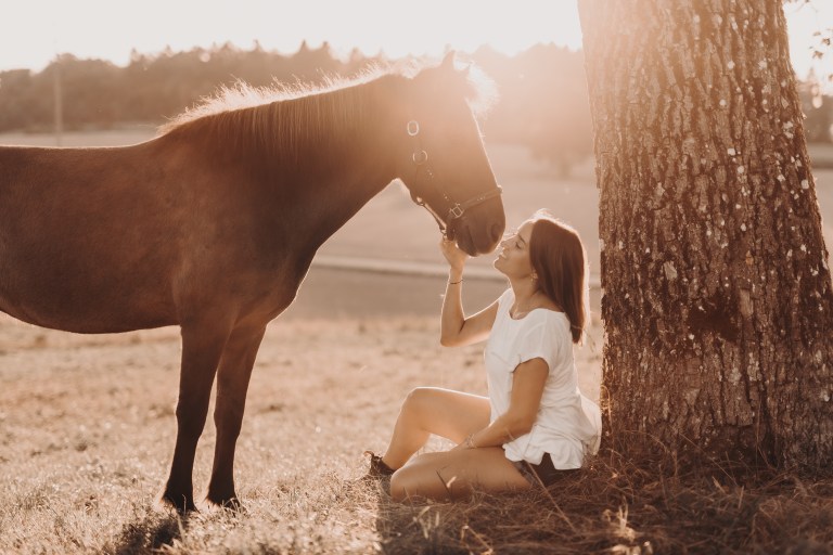 Eine Frau in einem weißen Hemd sitzt auf dem Boden, lehnt an einem Baum und berührt sanft das Gesicht eines braunen Pferdes, das neben ihr steht. Die untergehende Sonne wirft einen warmen, goldenen Schein über die friedliche Szene auf einer Wiese, ideal für Pferdefotografie.