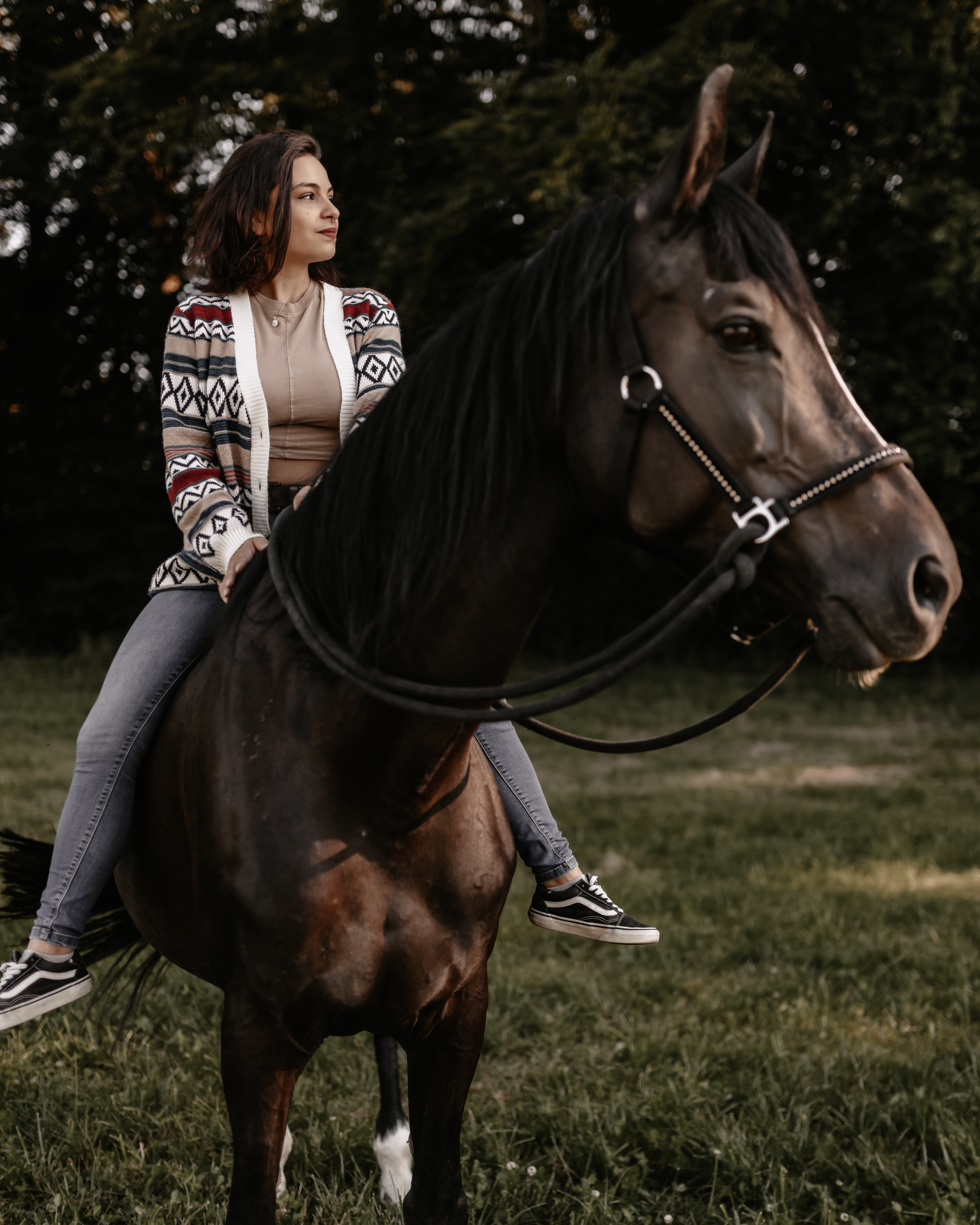 Eine Frau mit schulterlangem Haar, gemusterter Strickjacke und Jeans, reitet auf einem dunkelbraunen Pferd über eine Wiese. Mit ruhigem Gesichtsausdruck blickt die Fotografin in die Ferne und fängt den heiteren Moment ein, während Bäume den Hintergrund zieren.