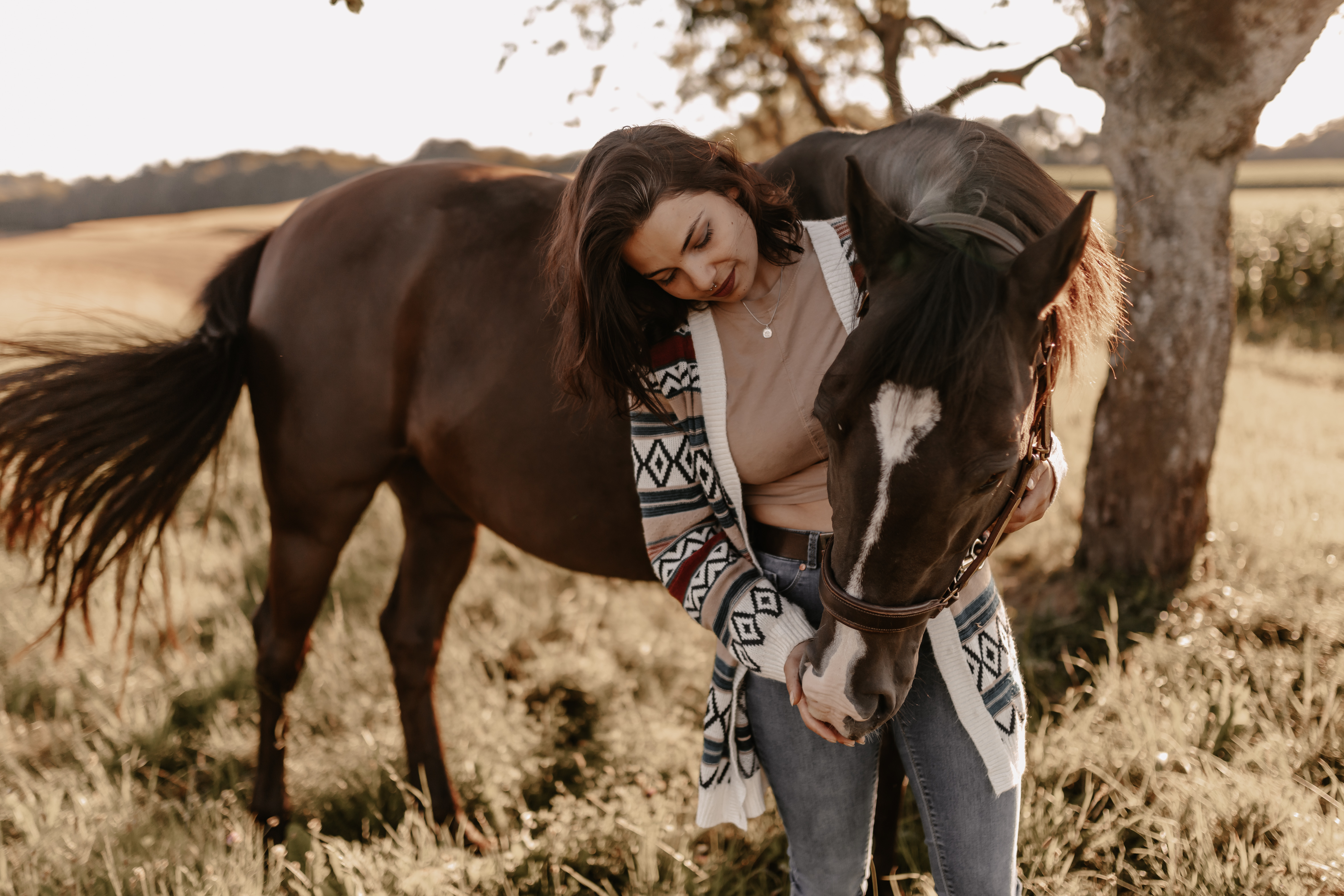 Eine Person mit langem dunklem Haar, die eine gemusterte Strickjacke und Jeans trägt, steht da und hält sanft den Kopf eines braunen Pferdes mit einem weißen Fleck auf der Stirn. Sie stehen draußen auf einer Grasfläche mit einem Baum und Feldern im Hintergrund und halten Momente für ein Tierfotoshooting eines erfahrenen Tierfotografen fest.