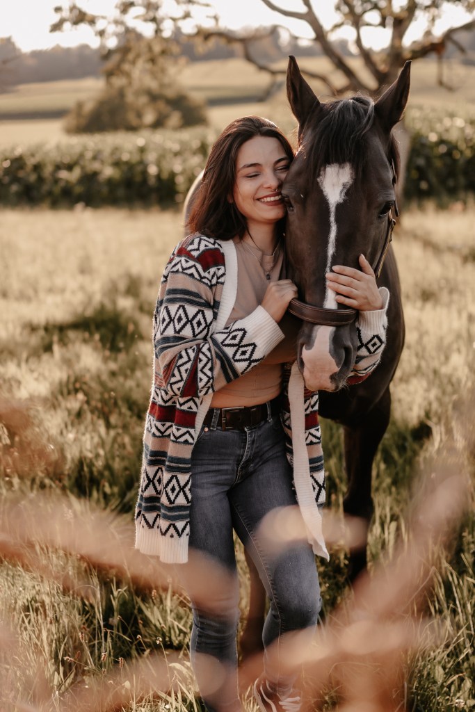 Eine lächelnde Frau in einer gemusterten Strickjacke und Jeans steht dicht neben einem dunklen Pferd mit einem weißen Streifen im Gesicht. Sie befinden sich auf einer Wiese, im Hintergrund fällt Sonnenlicht durch die Bäume, wunderschön eingefangen von einer erfahrenen Fotografin aus Aargau, die sich auf Tierfotografie spezialisiert hat.