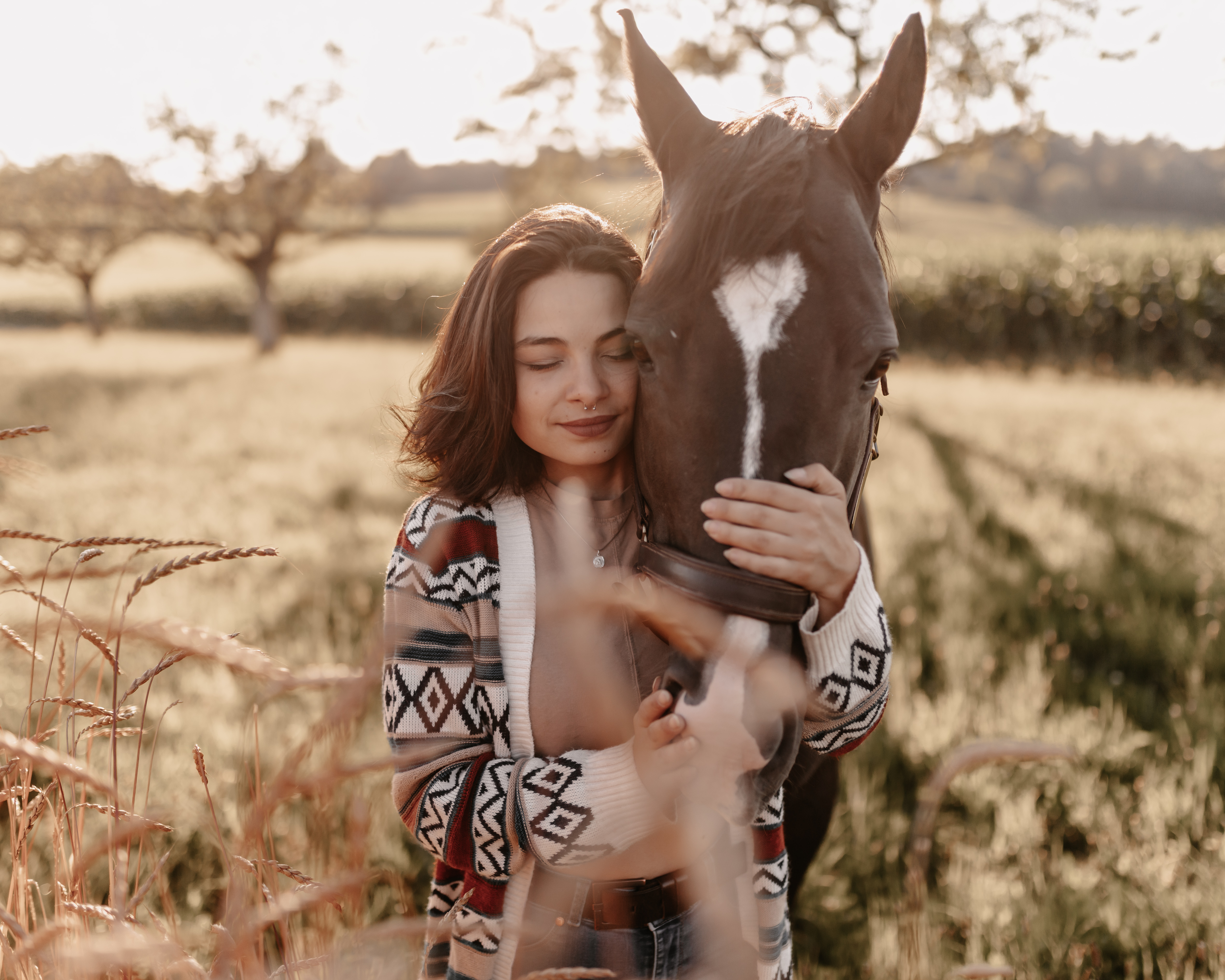 Eine Person mit schulterlangem dunklem Haar und einer gemusterten Strickjacke umarmt sanft ein braunes Pferd mit einer weißen Blesse im Gesicht. Sie stehen auf einer sonnenbeschienenen Wiese mit hohem Gras und Bäumen im Hintergrund, wunderschön aufgenommen von einer erfahrenen Fotografin, die sich auf Pferdefotografie im Aargau spezialisiert hat.
