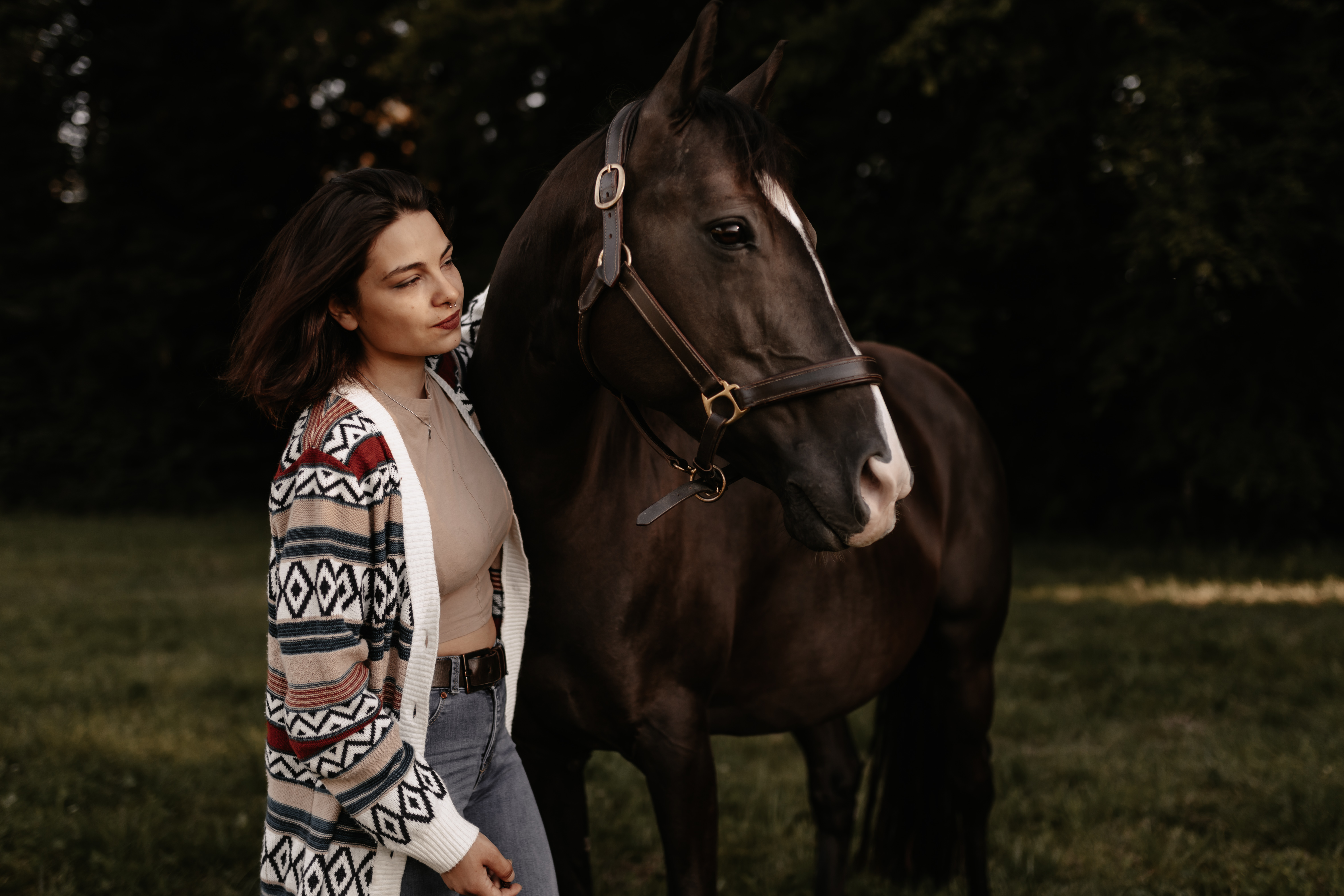 Eine Frau in einer gemusterten Strickjacke steht neben einem braunen Pferd auf einer Wiese mit Bäumen im Hintergrund, wahrscheinlich irgendwo im Aargau. Sie hält das Zaumzeug des Pferdes während eines Fotoshootings, das wie ein Tierfotograf aussieht, sanft und blickt dabei nachdenklich in die Ferne. Das Pferd ist ruhig und hat einen weißen Streifen im Gesicht.