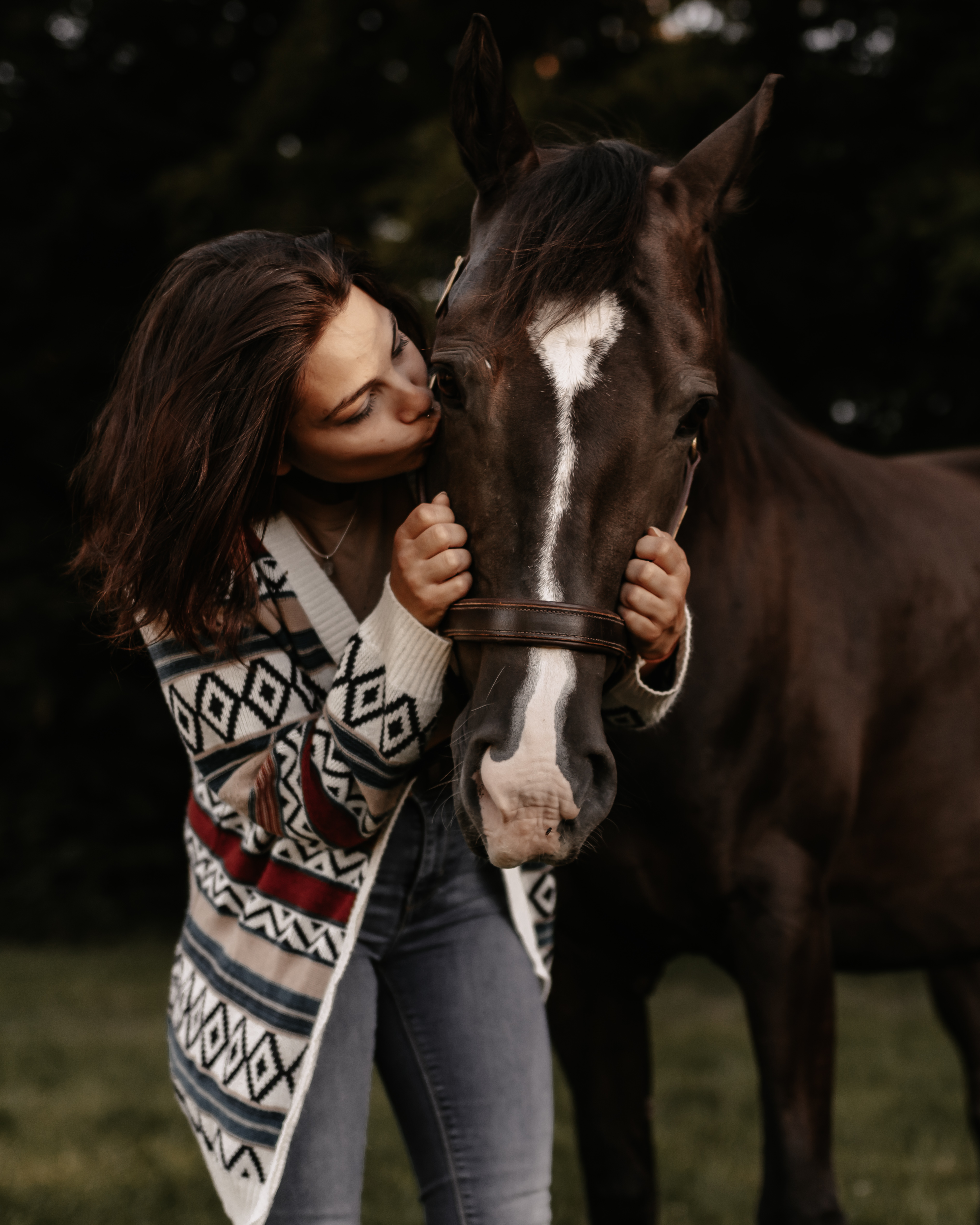Eine Frau in einem gemusterten Pullover und Jeans steht auf einer Wiese und küsst zärtlich ein braunes Pferd auf die Stirn. Das Pferd hat einen weißen Streifen im Gesicht und trägt ein Zaumzeug. Dieses Fotoshooting im Aargau wurde von einem erfahrenen Pferdefotografen aufgenommen und zeichnet sich durch einen dunklen, verschwommenen Laubhintergrund aus.
