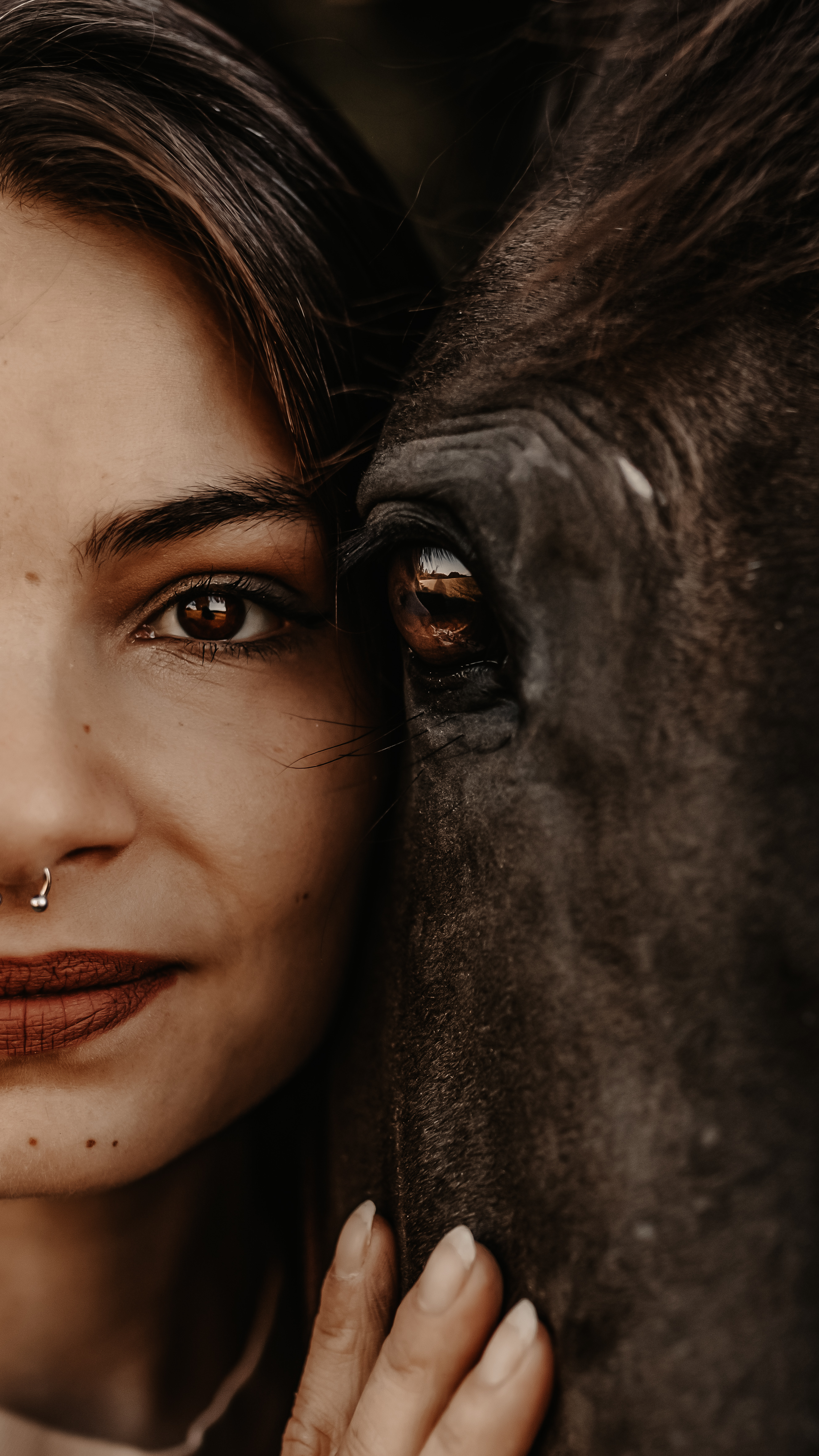 Eine Nahaufnahme zeigt die Gesichter einer Frau und eines Pferdes nebeneinander während eines ruhigen Fotoshootings. Die Frau hat dunkle Haare, einen Nasenring und trägt Lippenstift. Ihre Hand ruht sanft auf dem Gesicht des Pferdes. Beide scheinen direkt in die Kamera zu blicken und fangen einen intimen Moment durch die Linse eines talentierten Tierfotografen ein.