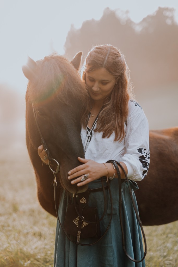 Eine Frau mit langen Haaren, die eine weiße Bluse und einen blaugrünen Rock trägt, streichelt sanft den Kopf eines braunen Pferdes. Sie stehen auf einem sonnenbeschienenen Feld mit einem weichen, dunstigen Hintergrund in einem verträumten Fantasy-Fotoshooting, das einen ruhigen und intimen Moment zwischen ihnen schafft.