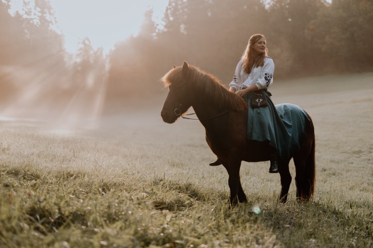 Eine Frau sitzt auf einem braunen Pferd auf einer sonnenbeschienenen Wiese mit taufrischem Gras. Sie trägt eine weiße Bluse und einen blauen Rock und blickt mit heiterem Gesichtsausdruck nach rechts. Hohe Bäume im Hintergrund werden sanft von der frühen Morgensonne beleuchtet, als würden sie die Essenz tierfotografischer Kunst einfangen.