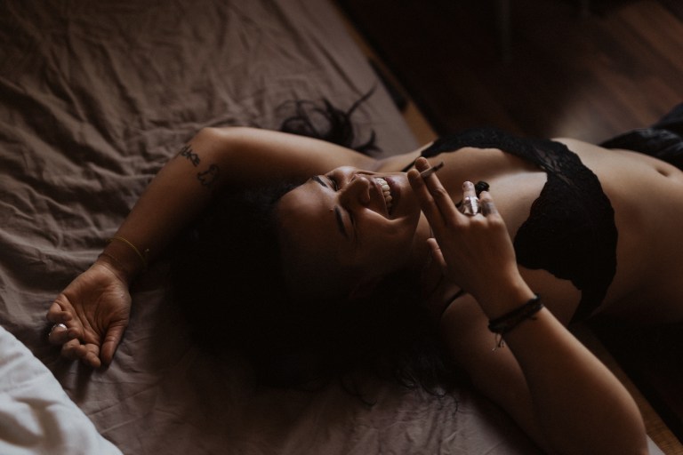 Eine Person mit dunklem Haar und schwarzer Unterwäsche liegt lächelnd auf einem Bett und hält eine Zigarette in der Hand. Ihr rechter Arm ist über dem Kopf ausgestreckt und zeigt Tätowierungen, während ihr linker Arm angewinkelt ist und an ihren Fingern ein paar Ringe zu sehen sind. Die Beleuchtung ist schwach und warm und fängt den Moment in einem natürlichen Porträtstil einer talentierten Fotografin ein.